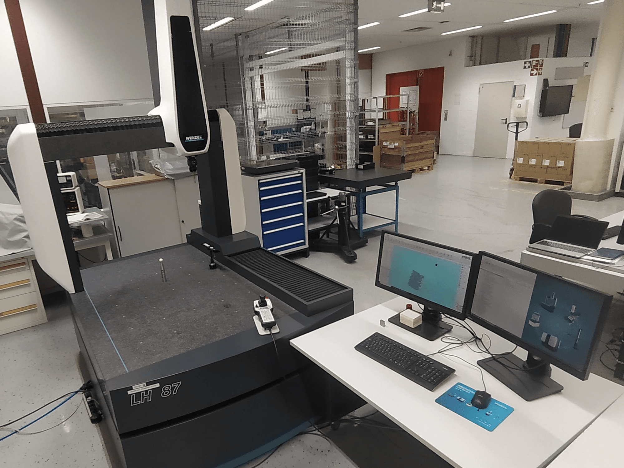 Souřadnicový měřicí stroj Wenzel LH 87 viditelný v laboratoři, se dvěma monitory a měřicí sondou na stole.