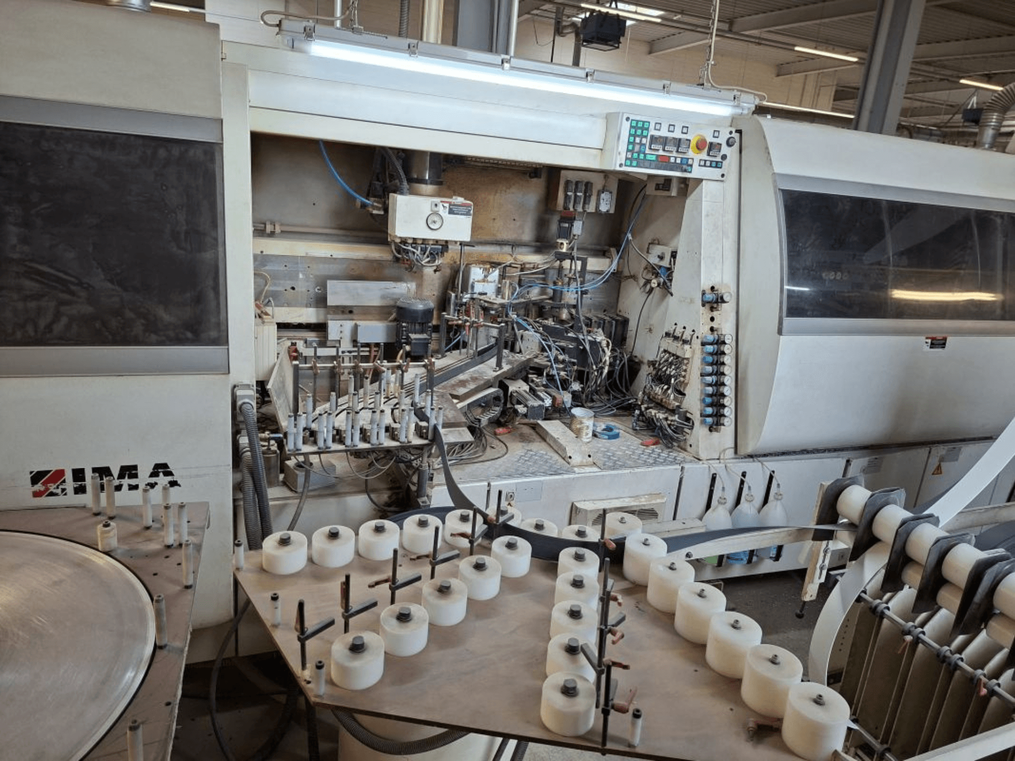 Strojní zařízení IMA při pohledu z boku, kde jsou vidět složité mechanické součásti a kulatý stůl s bílými cívkami.