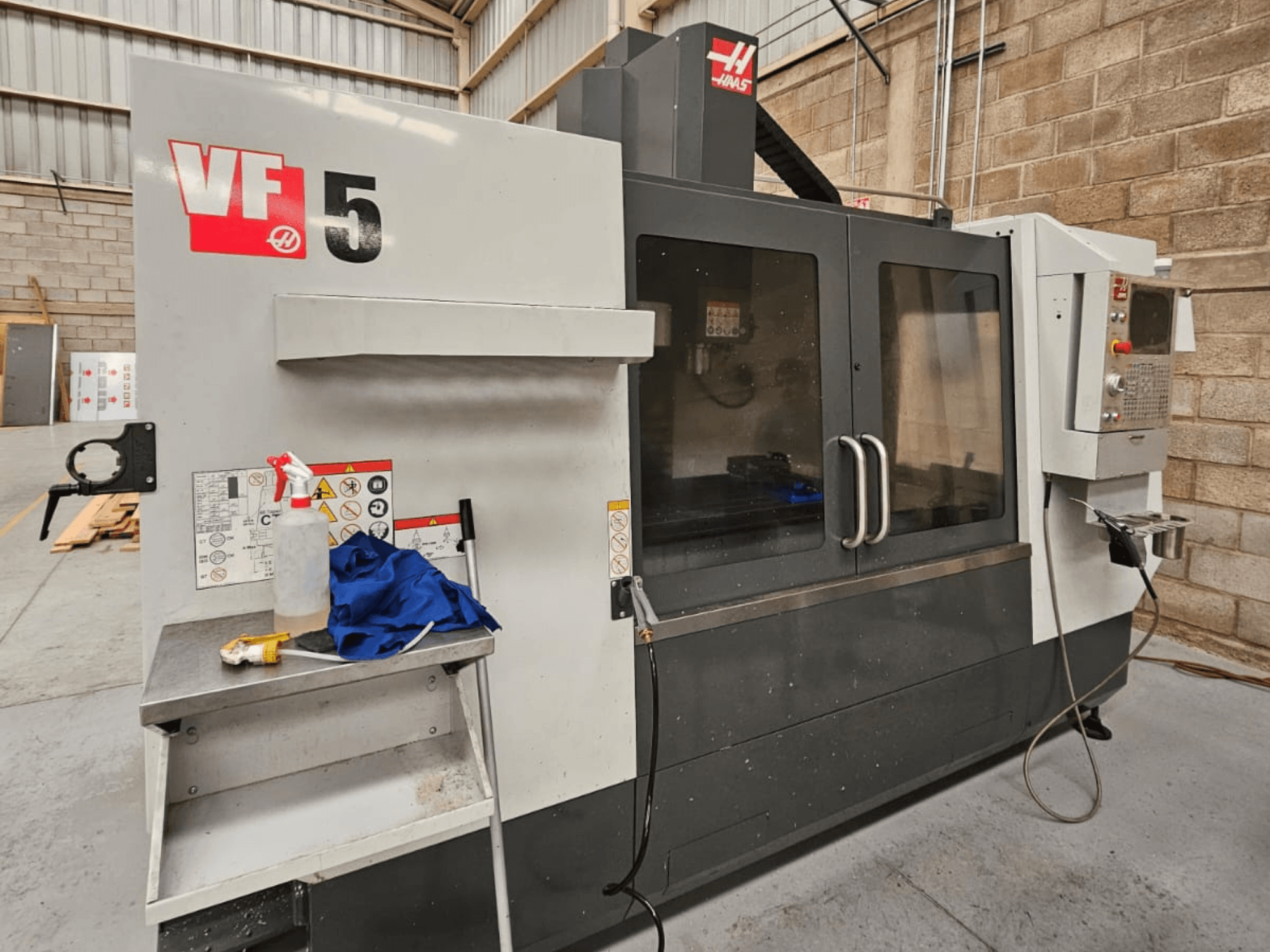 CNC frézka Haas VF-5 v čelním pohledu, s ovládacím panelem, lahví s chladicí kapalinou, modrou látkou a bezpečnostními pokyny.