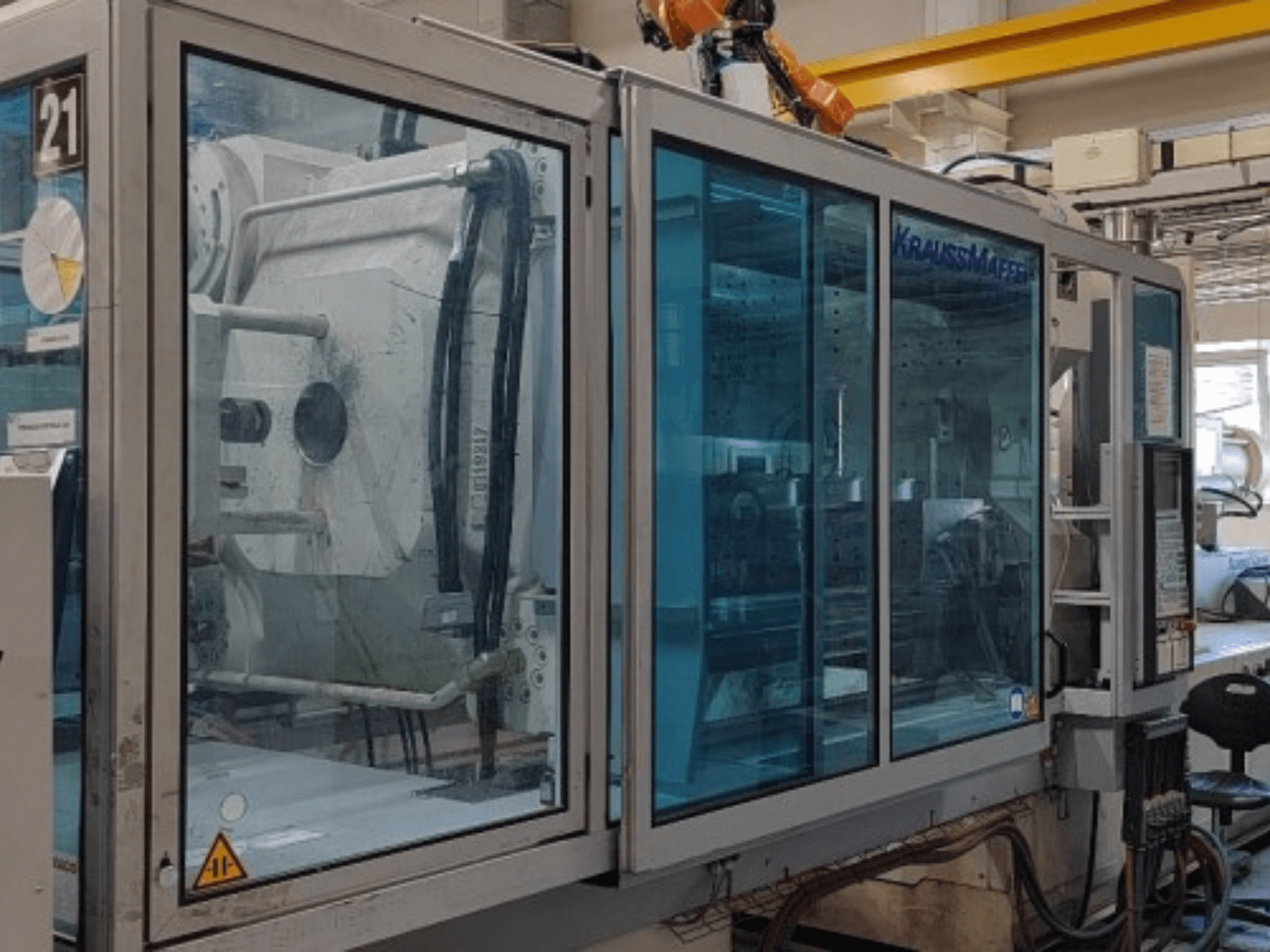Hydraulický vstřikovací lis Krauss Maffei KM 575-2700 C2, boční pohled, s průhlednými bezpečnostními panely a ovládacím panelem.