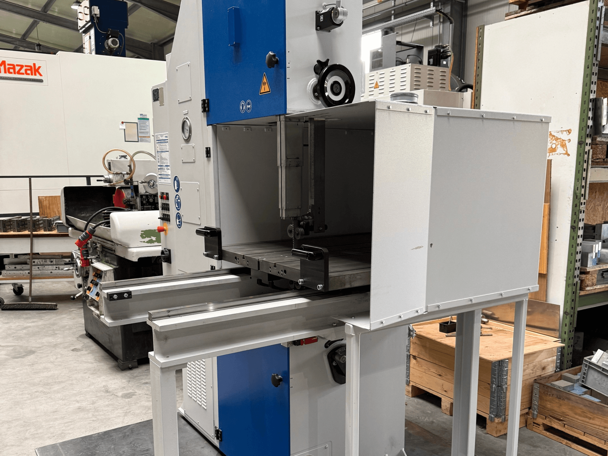 Průmyslový stroj Mazak při pohledu zepředu, s modrým ovládacím panelem, kovovým stolem a bezpečnostními upozorněními.