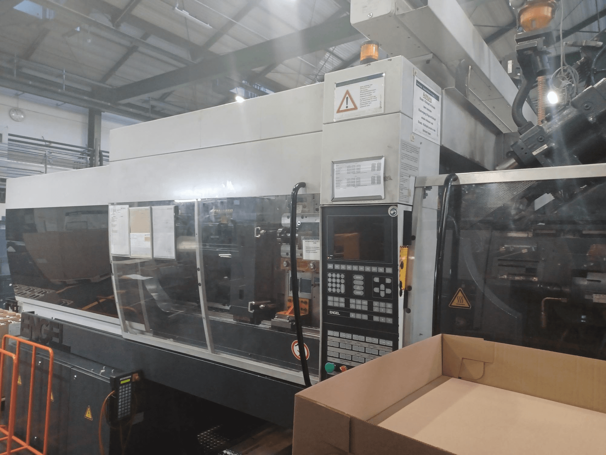 Vstřikovací stroj Engel, pohled zepředu, s ovládacím panelem, bezpečnostními štítky a prázdnou krabicí umístěnou poblíž.