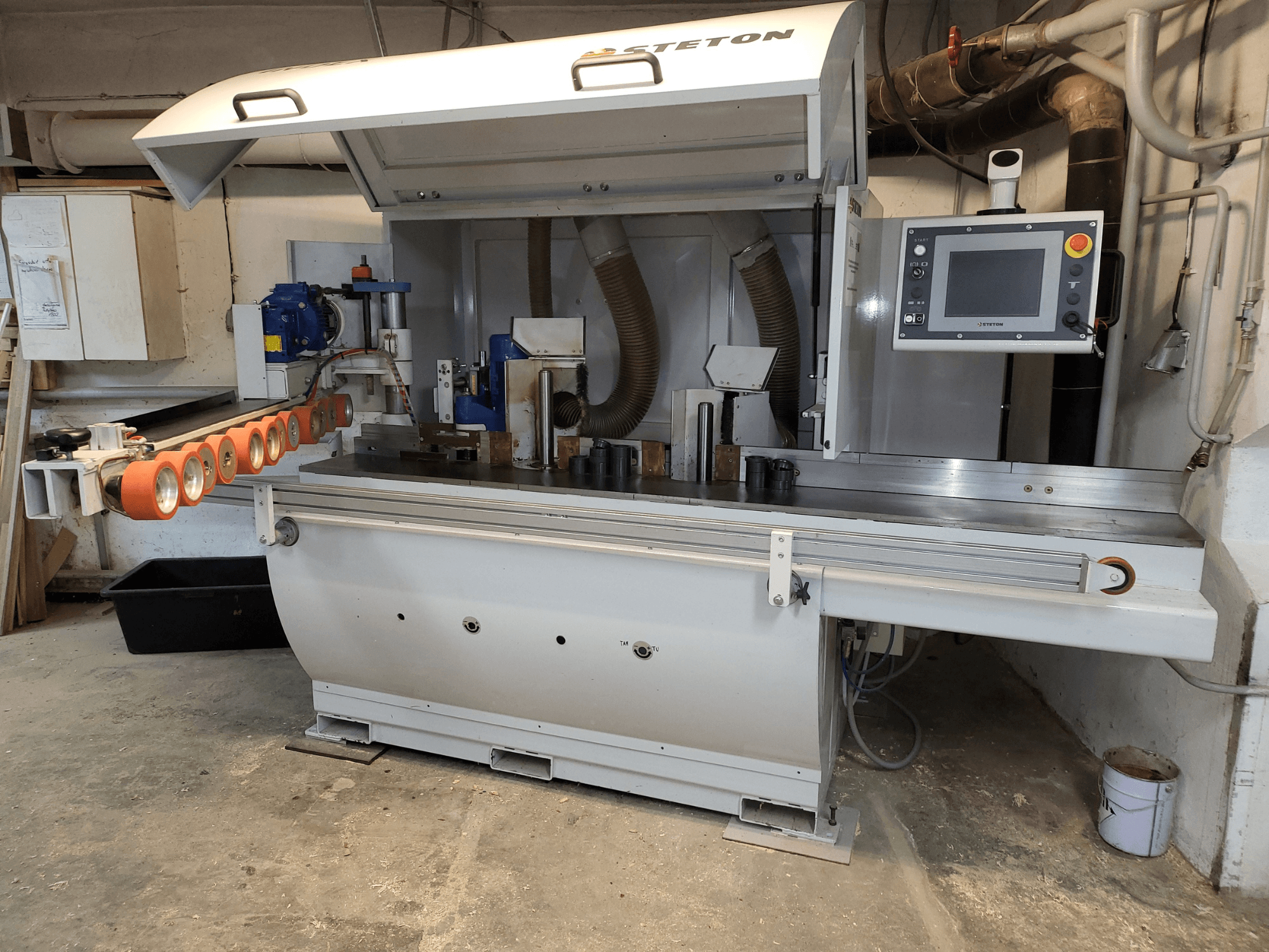 Pohled zepředu na dřevoobráběcí stroj Stetton s ovládacím panelem, podávacími trubkami a různými kusy dřeva na stole.