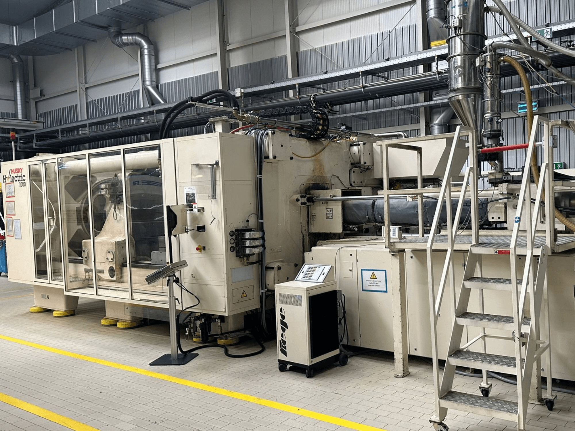Pohled zepředu na elektrický vstřikovací lis Husky Hylelectric H1000 RS95/80 v továrním prostředí, s ovládacím panelem a schůdky.