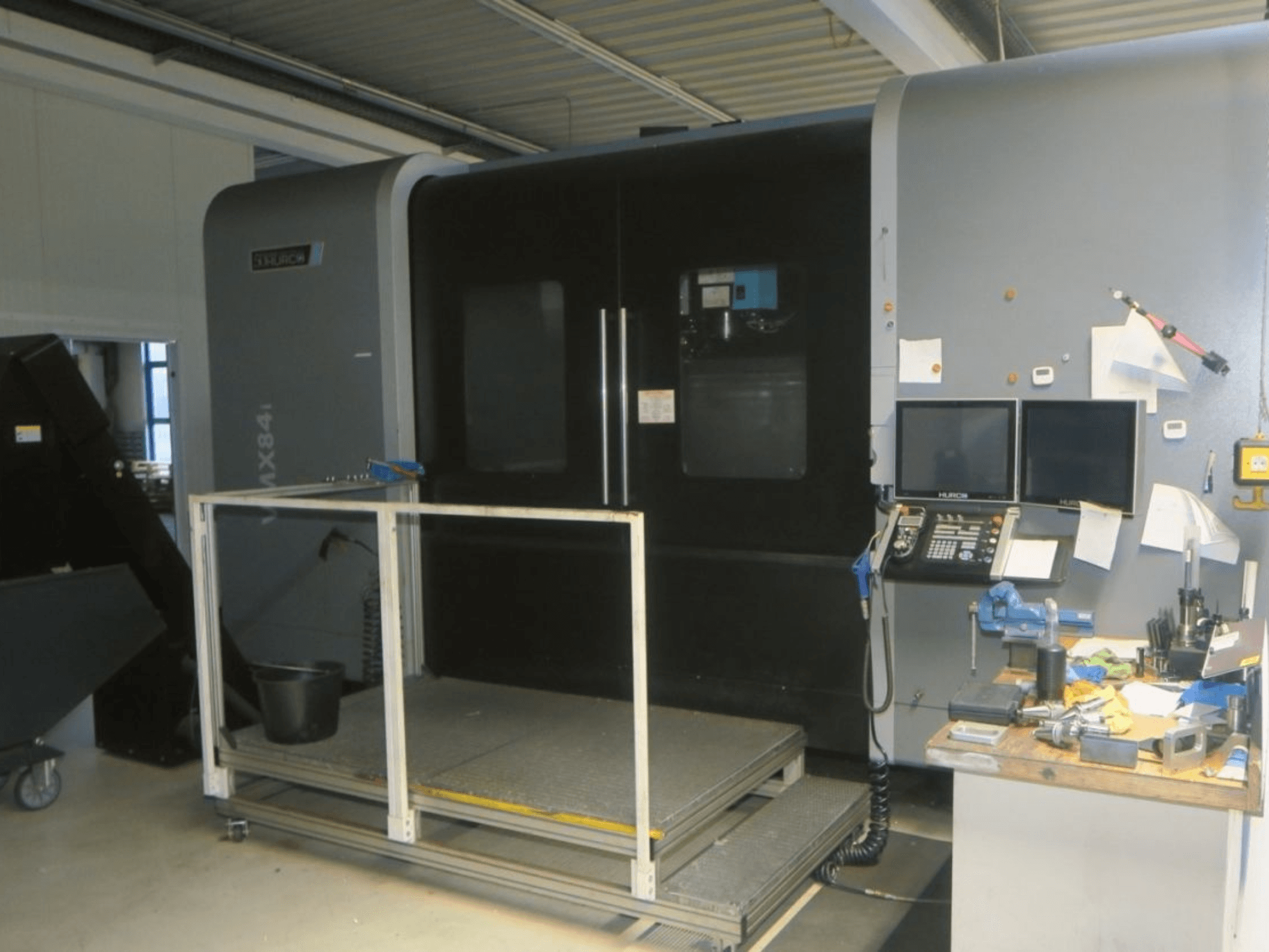 CNC stroj Hurco MX84 v čelním pohledu, zobrazující dva monitory, ovládací panel a pracovní prostor s různými nástroji.