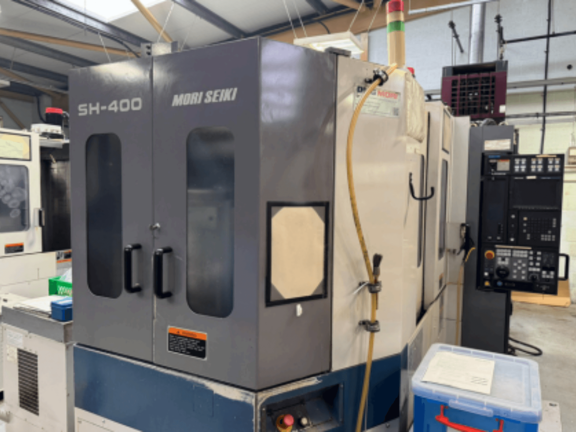 Stroj Mori Seiki SH-400 CNC, pohled zepředu, se dvěma dveřmi, ovládacím panelem a přípojnou hadicí. Modrý úložný kontejner poblíž.