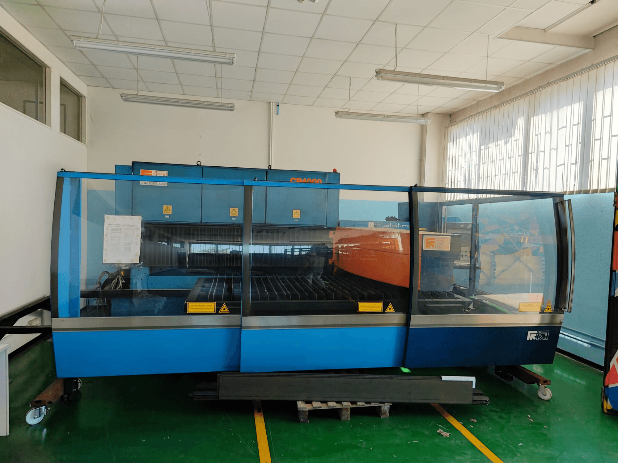 Pohled zepředu na laserový řezací stroj CONVERGENT LASERS CP4000 s CO₂ laserem v modro-oranžovém provedení, v dobře osvětlené dílně.