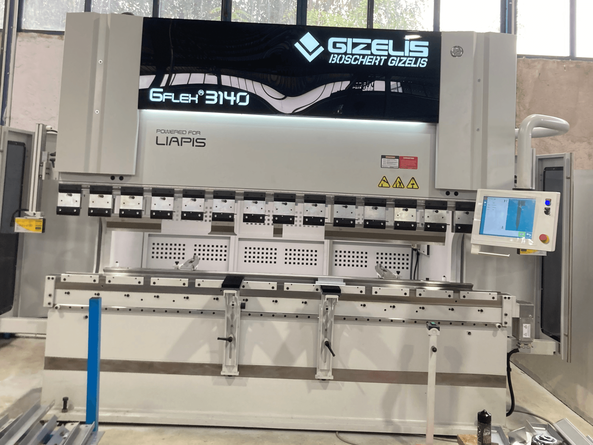 Hydraulický lis GIZELIS G-FLEX 3140 v čelním pohledu s ovládacím displejem, s několika ohýbacími nástroji a bezpečnostními štítky.