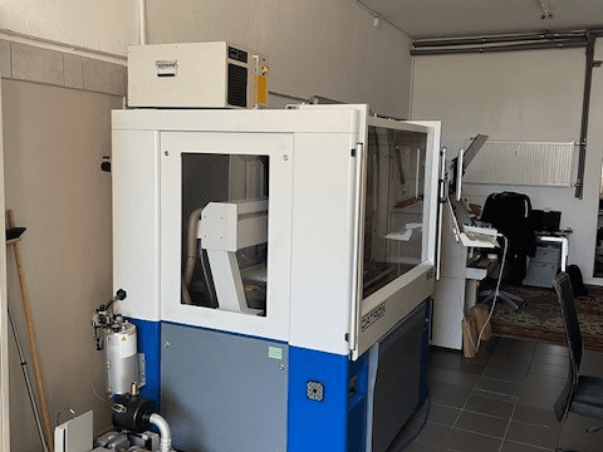 Modrobílý průmyslový stroj s průhlednými panely, pohled zepředu, včetně přístupu k ovládání a viditelné ventilační jednotky.