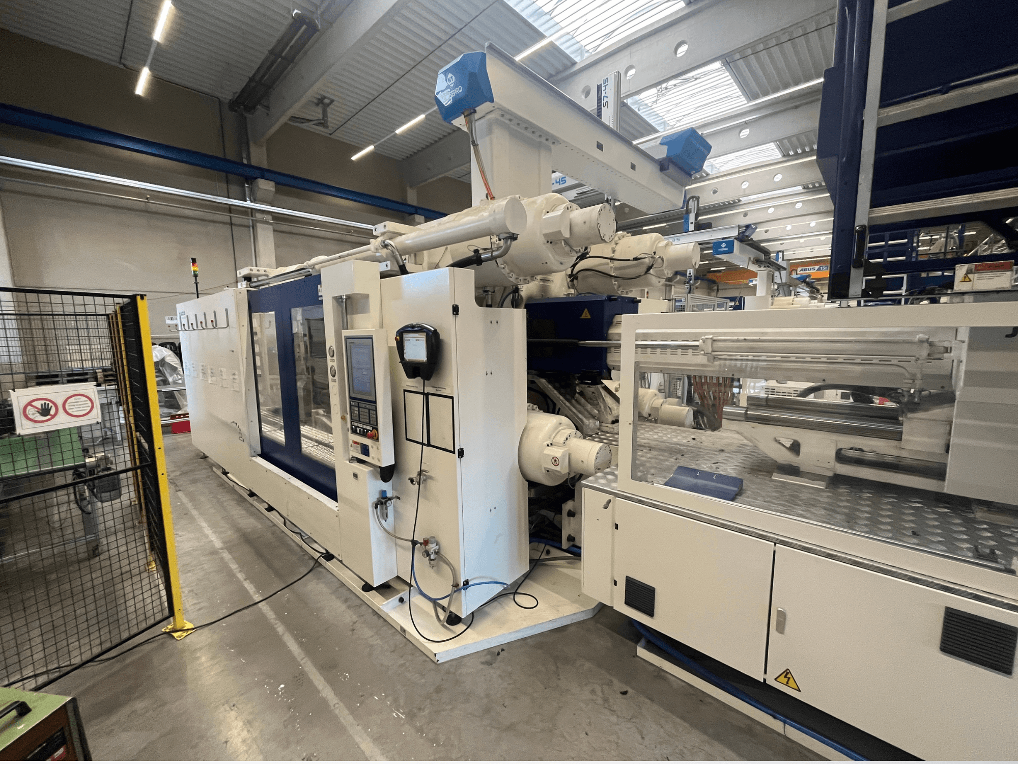 Vstřikovací stroj v bílé a modré barvě, pohled zepředu, s ovládacím panelem, robotickým ramenem a bezpečnostním značením.