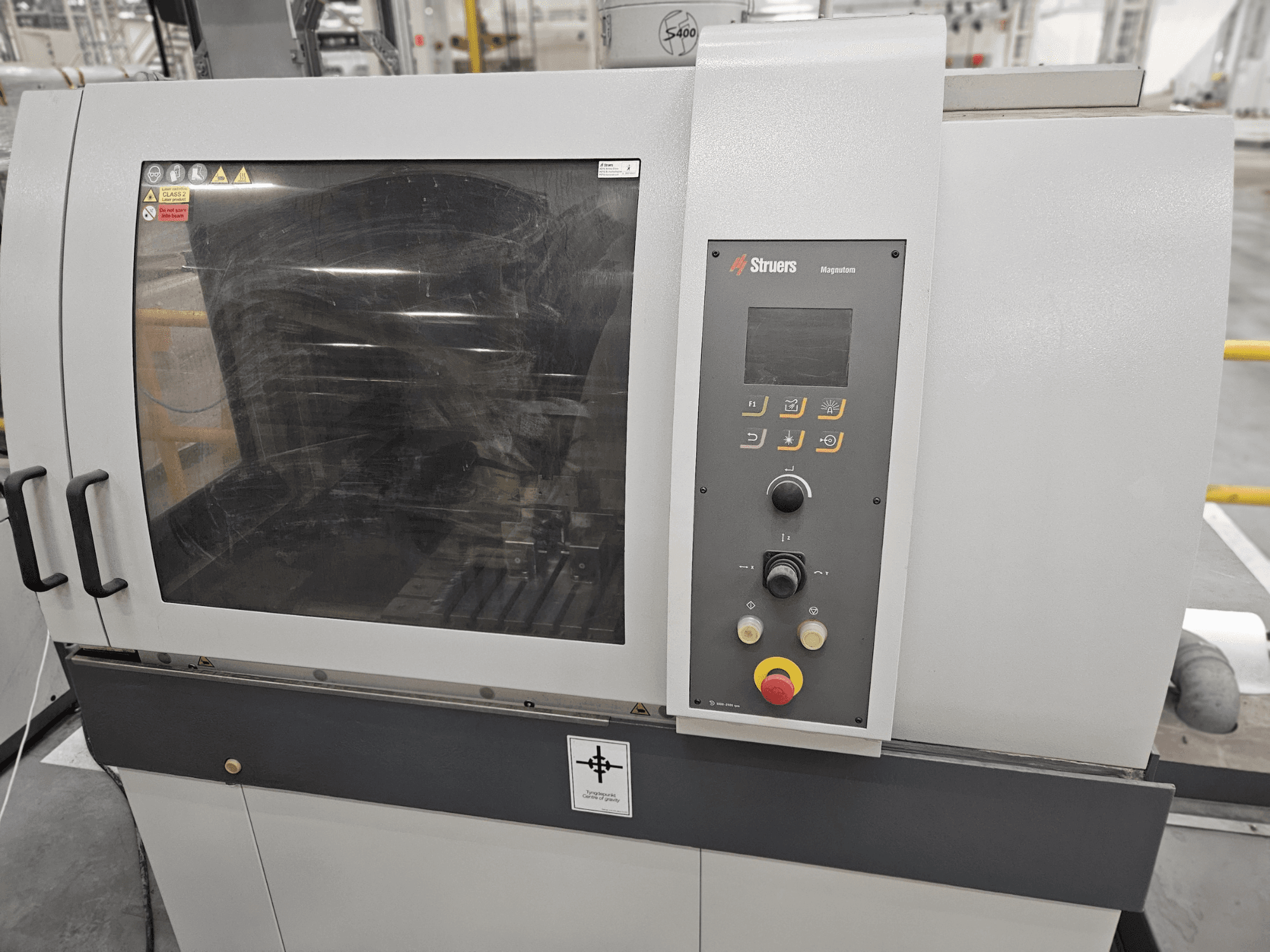 Pohled na stroj Struers Magpentor zepředu s ovládacím panelem, tlačítky a velkým průzorem. Viditelné technické štítky.