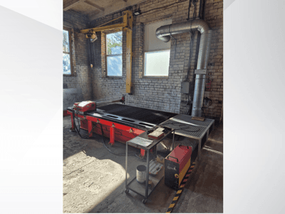 Červený CNC plazmový řezací stůl s ovládacím panelem a kovovým výfukovým potrubím, pohled z boku v dobře osvětlené dílně.