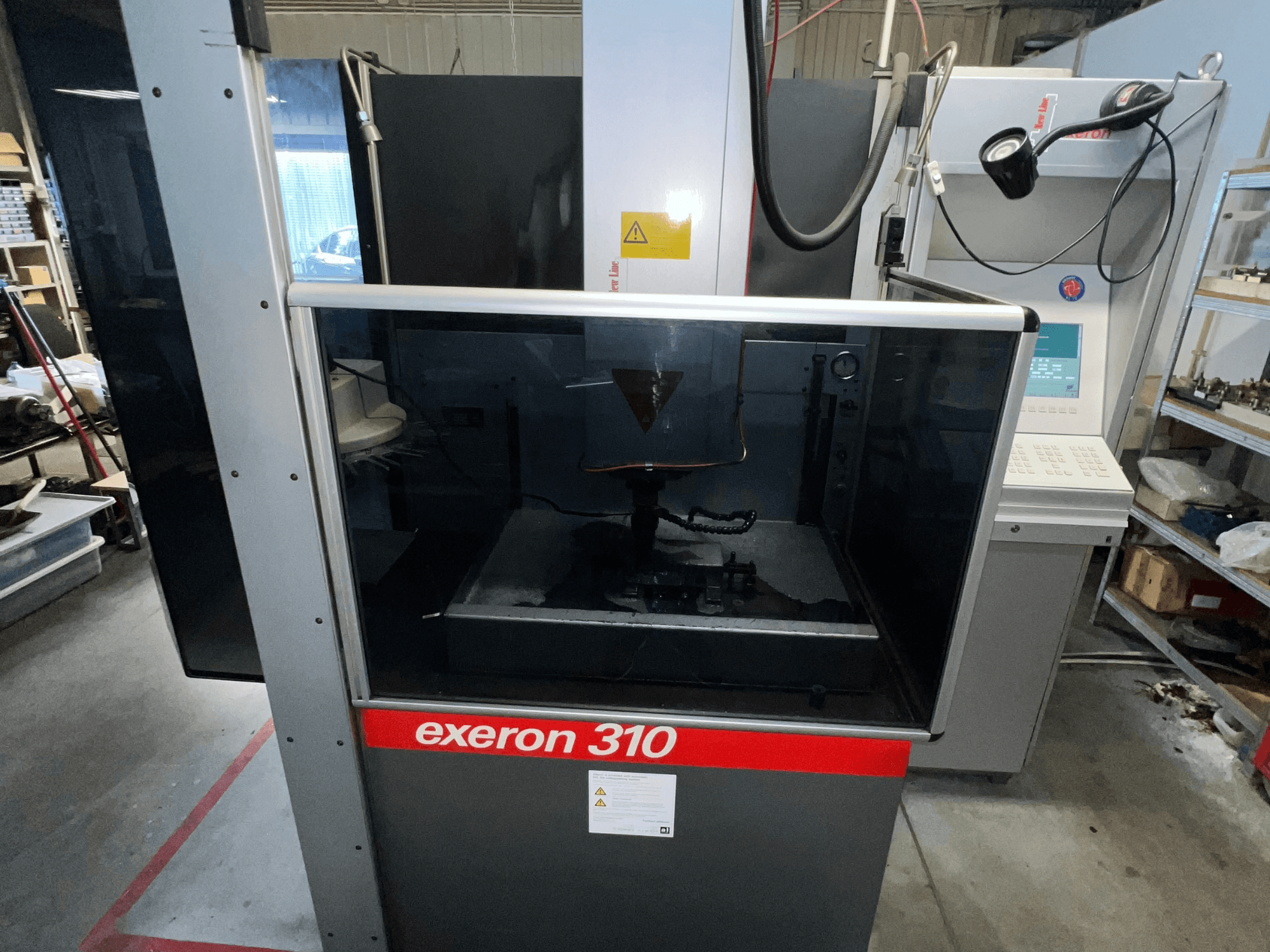 Pohled zepředu na stroj Exeron 310, který ukazuje skleněnou komoru a ovládací panel vedle polic na pracovišti.