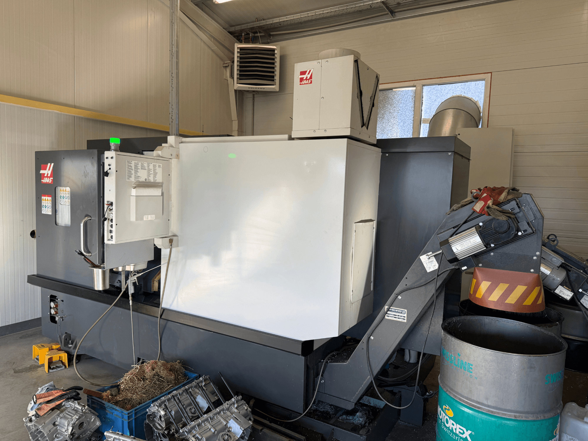 CNC stroj Haas s ovládacím panelem, boční pohled; viditelné vedení chladicí kapaliny a oleje; kovový odpad v modrém zásobníku; průmyslové prostředí.