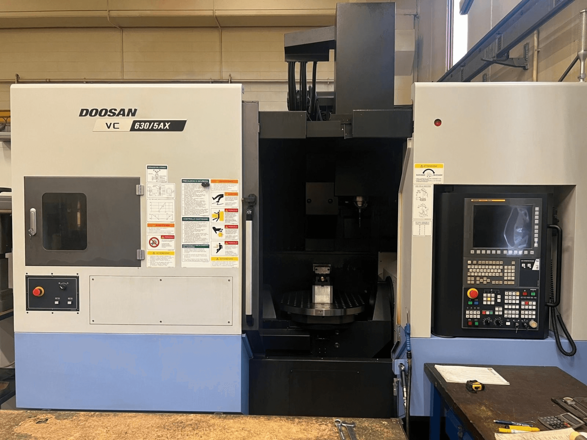 Pohled zepředu na vertikální obráběcí centrum DOOSAN VC 630/5AX, na kterém je vidět ovládací panel a oblast vřetena s viditelnými bezpečnostními štítky.