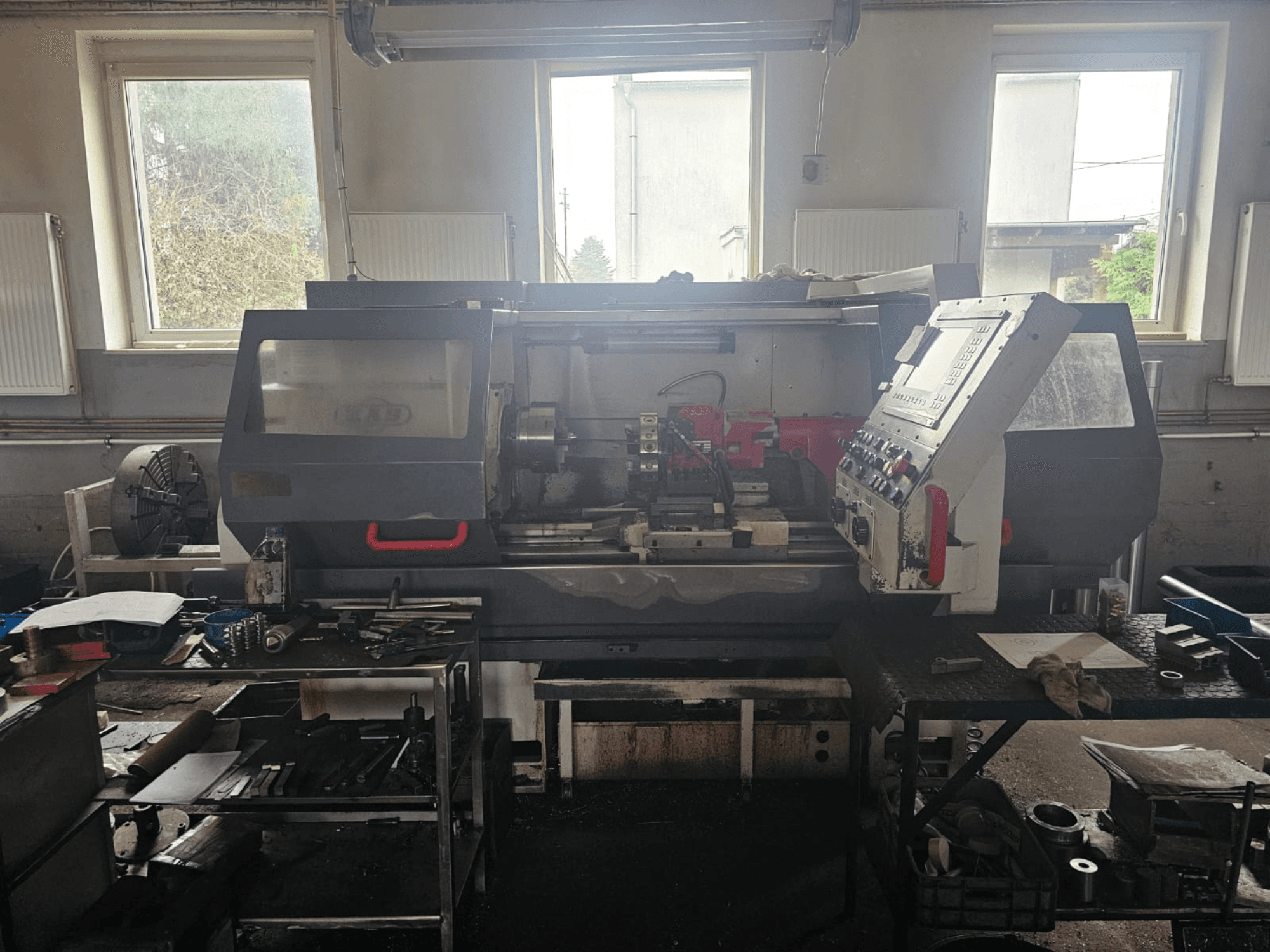 Soustruh v dílně, pohled zepředu, s ovládacím panelem a nástroji na stolech, obklopený okny a přeplněným pracovním prostorem.