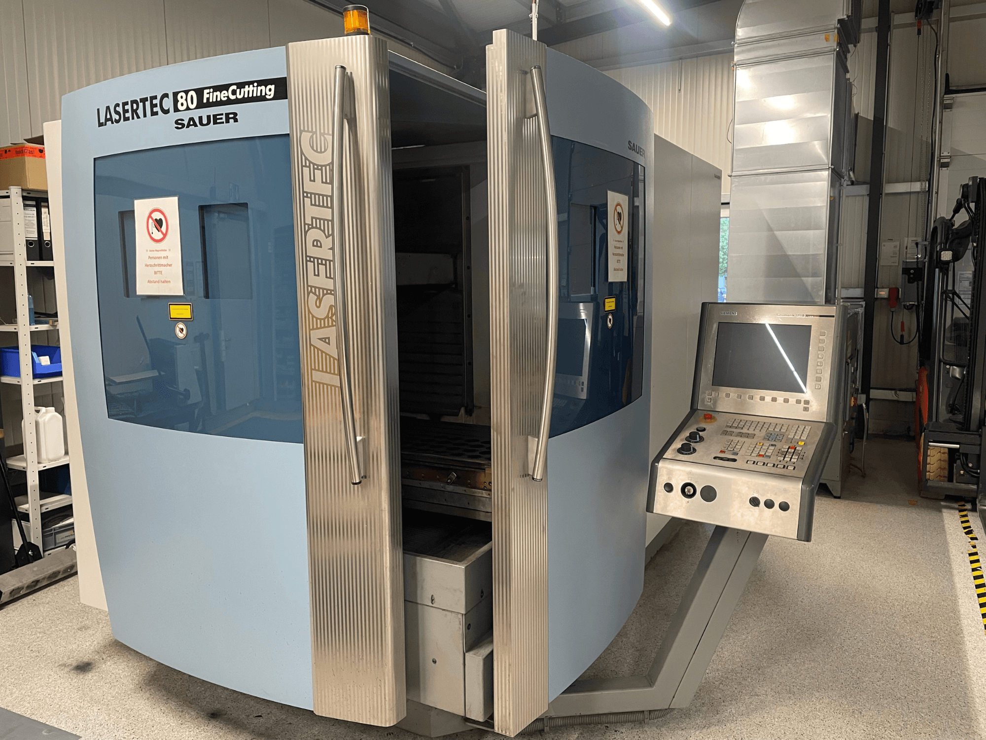 Pohled zepředu na stroj na řezání plechu DMG Sauer Lasertec80 FineCutting s ovládacím panelem, otevřenými dvířky a viditelným bezpečnostním značením.