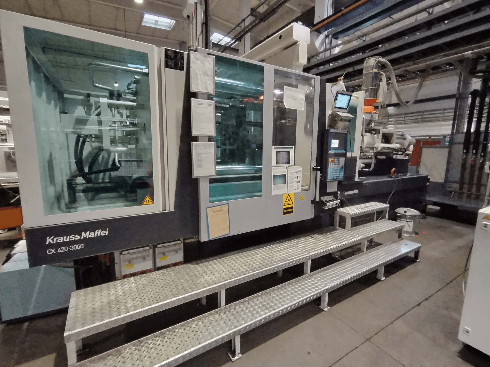 Vstřikovací stroj Krauss Maffei CX 420-3000, boční pohled, s viditelným ovládacím panelem a bezpečnostním značením, umístěný na kovových stupních.