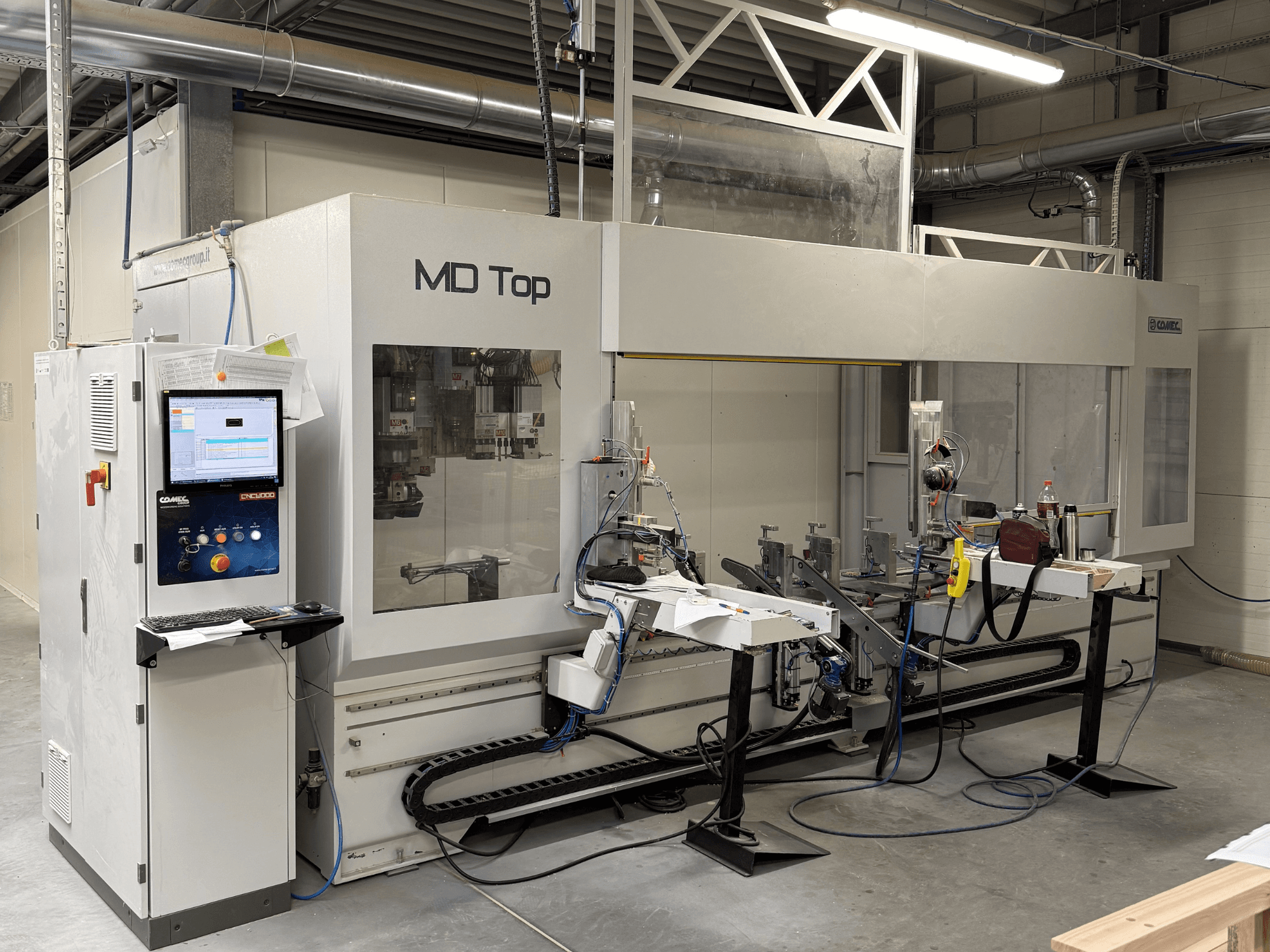 Stroj MD Top CNC při pohledu zepředu, s ovládacím panelem, robotickými rameny a pracovním prostorem s nástroji a materiály.
