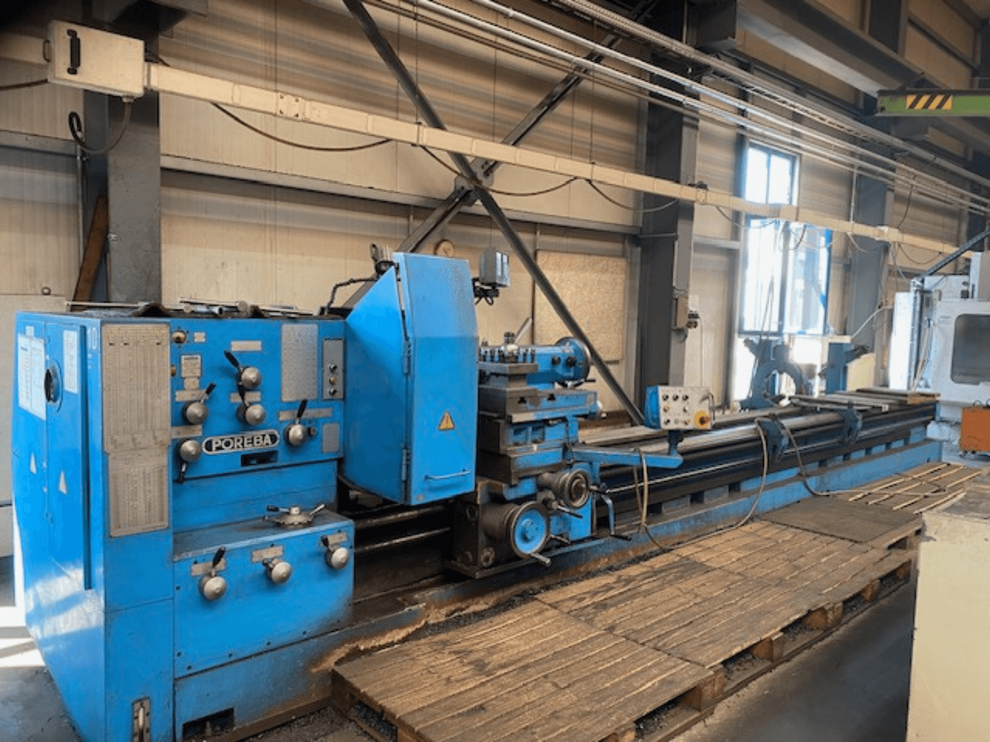 Pohled zepředu na modrý soustruh POREBA s ovládacími knoflíky, opěrkou nástrojů a dřevěnými paletami pod nimi.