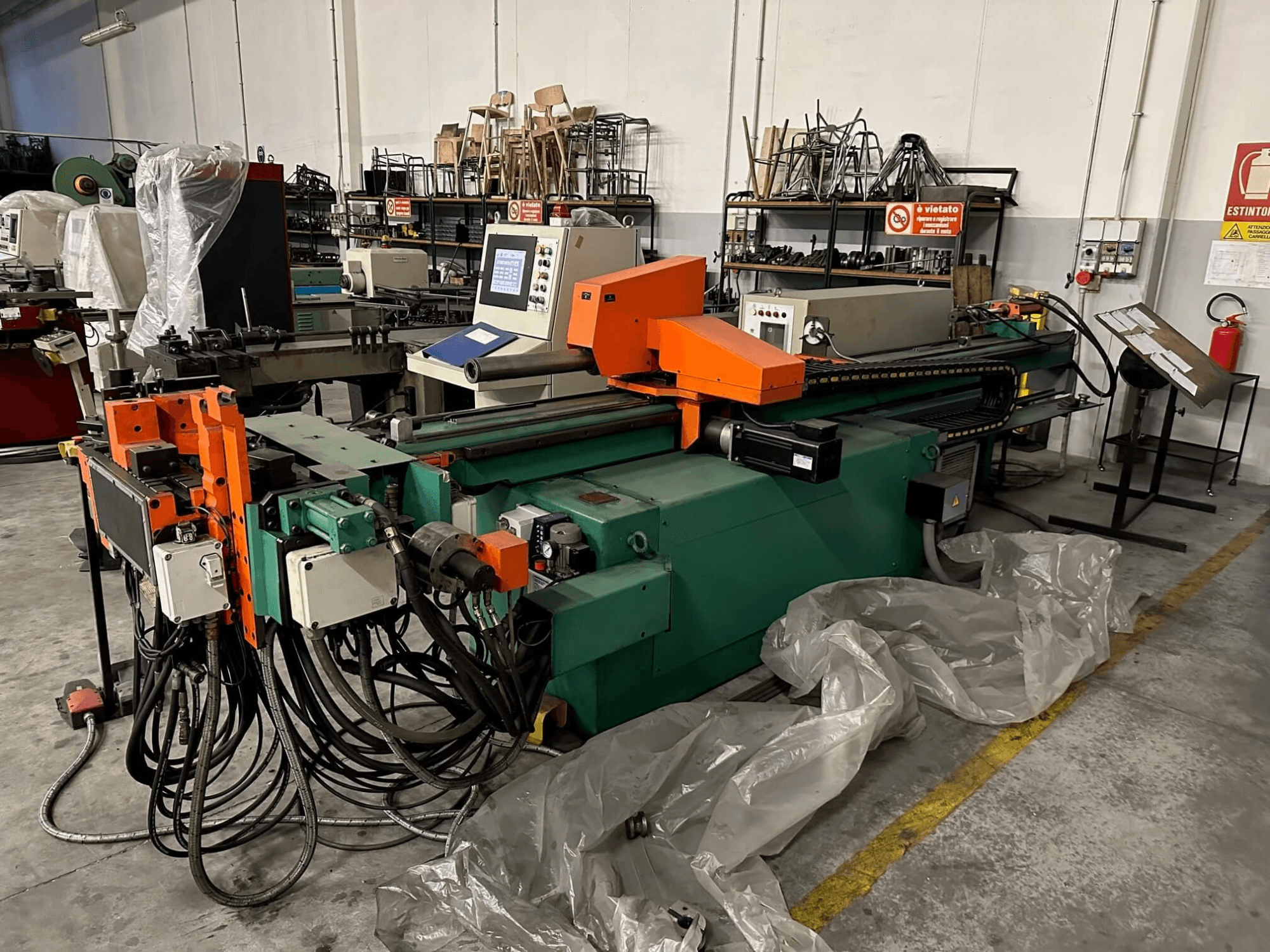 Průmyslový stroj s oranžovo-zelenou karoserií, ovládacím panelem a četnými kabely, pohled zepředu v dílně.