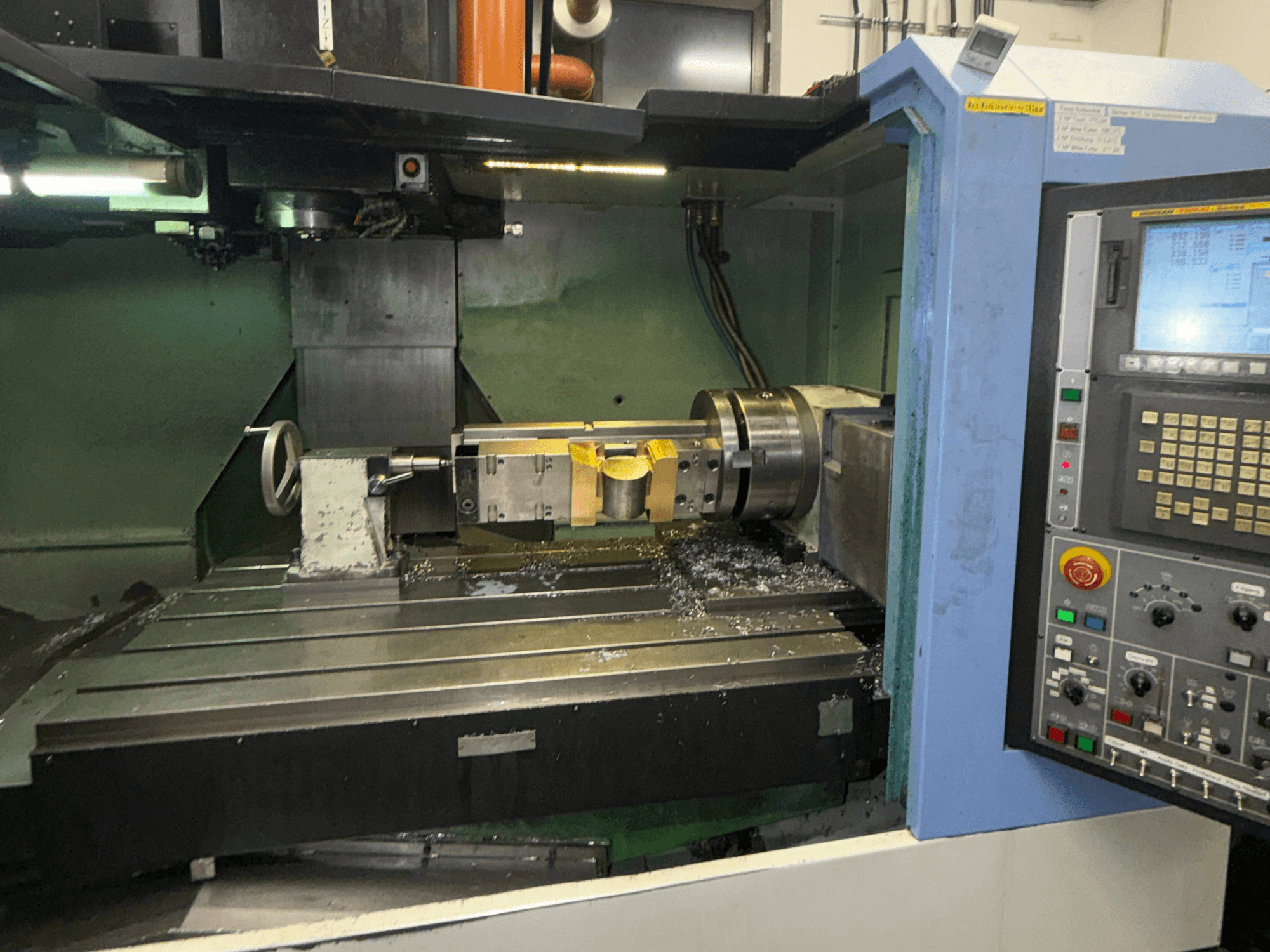 Vertikální obráběcí centrum DOOSAN DNM650, pohled zepředu, na kterém je vidět řezací nástroj a ovládací panel, na stole jsou vidět kovové třísky.