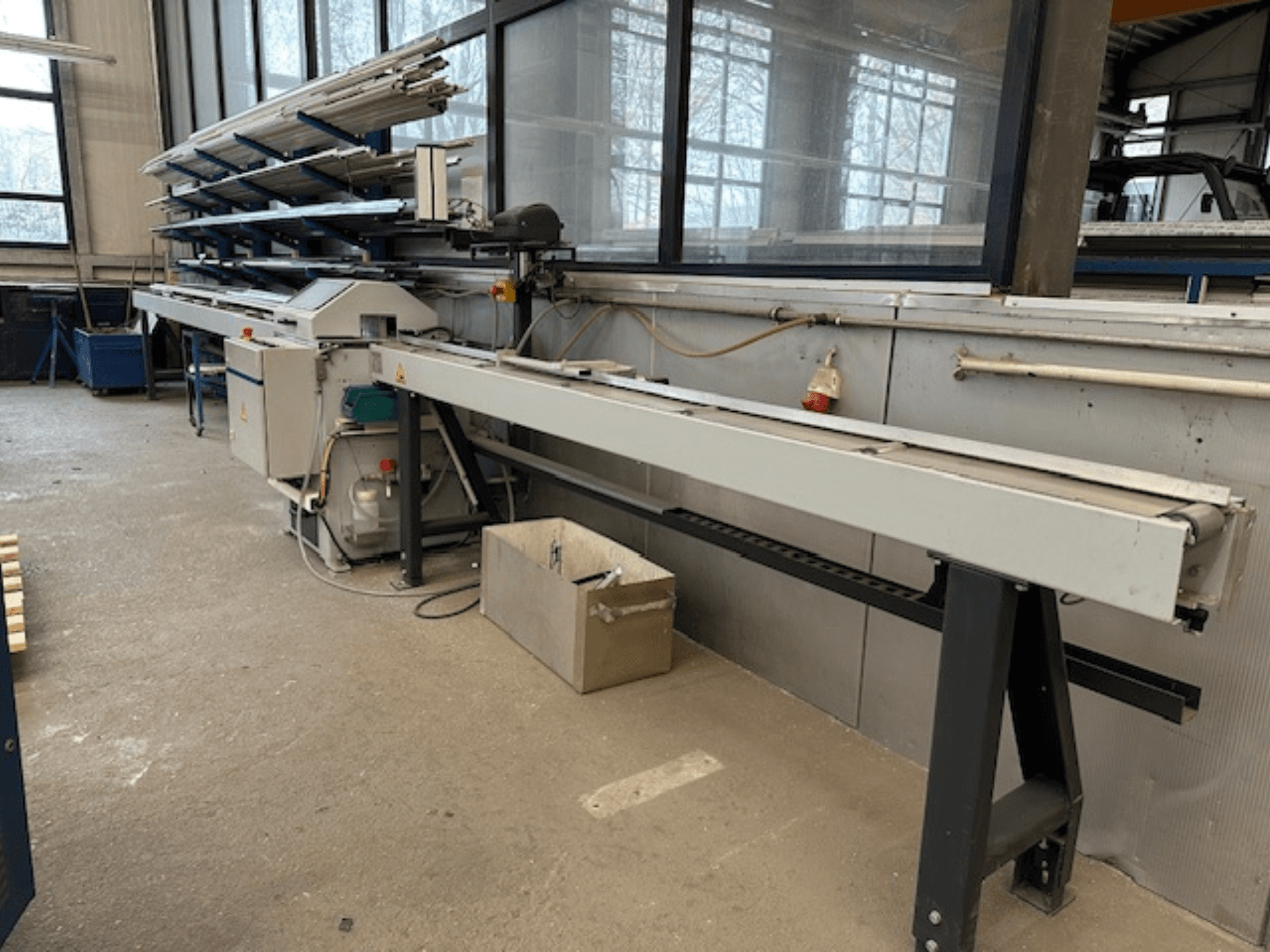 Kotoučová pila na kov SCHÜCO PGS v dílně, boční pohled s nastavitelným řezacím ložem a policí s kovovými profily nad ním.