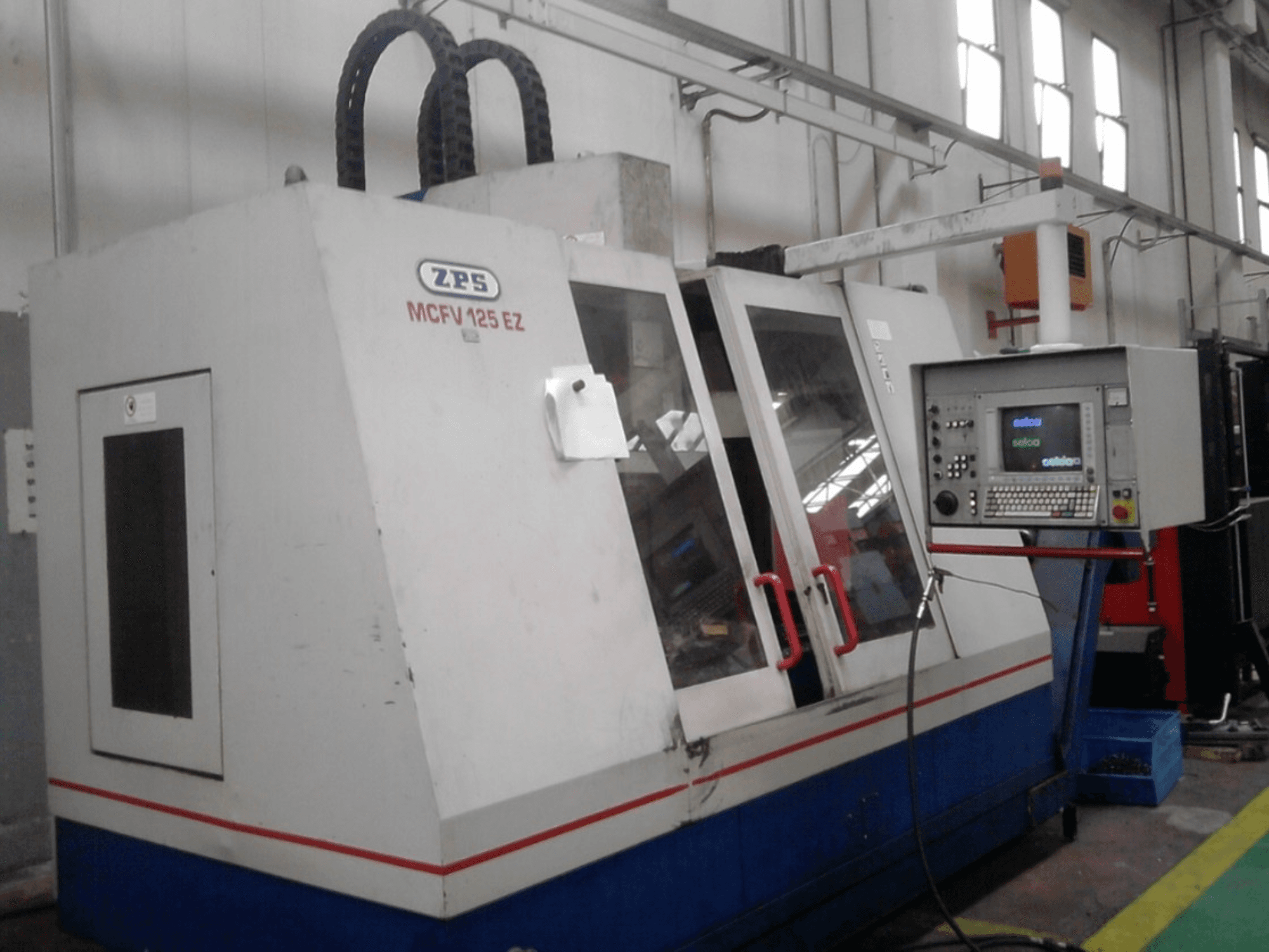 Stroj ZPS MCFV 125 EZ CNC, pohled zepředu s ovládacím panelem a červenými rukojeťmi, v dílně s viditelnými hadicemi.