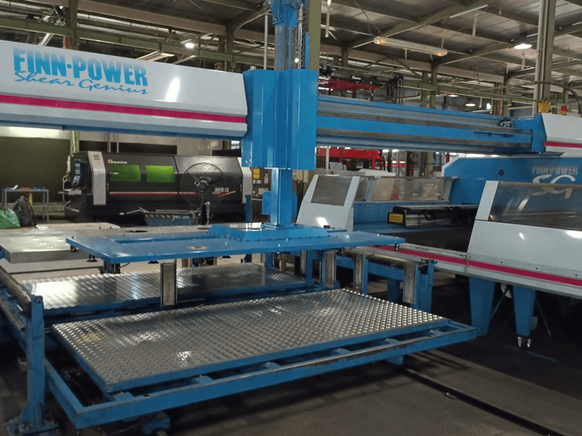 CNC děrovací stroj Finn-Power SG6 „Shear Genius“ z čelního pohledu, na kterém jsou vidět kovové stoly a konstrukční prvky.