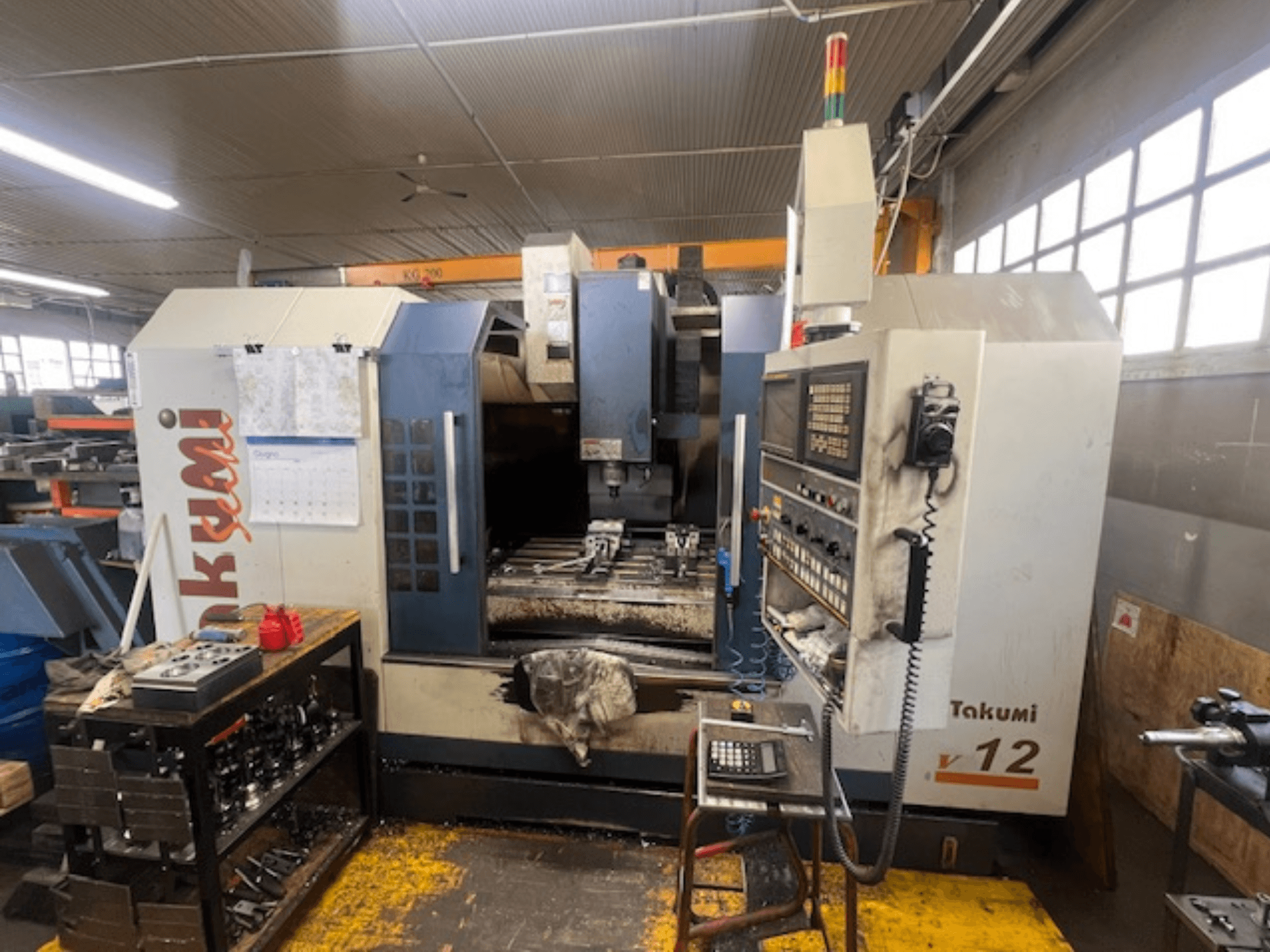 CNC frézka Takumi V12 v bočním pohledu, zobrazující ovládací panel, řezný prostor a nástroje na pracovní stanici.