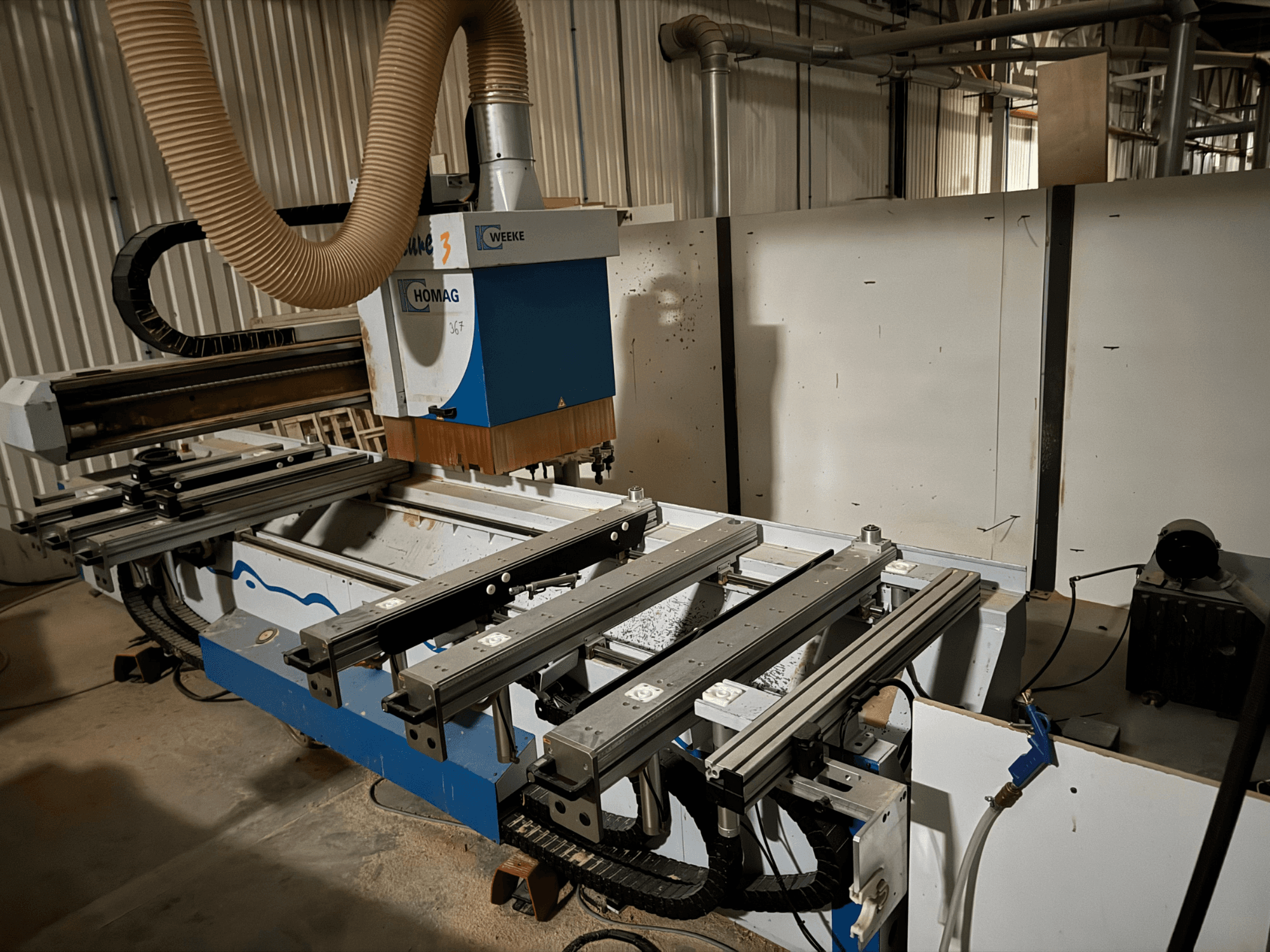 CNC stroj HOMAG v modrobílém provedení, pohled z boku, s posuvnými lištami, systémem odsávání prachu a namontovanými nástroji.