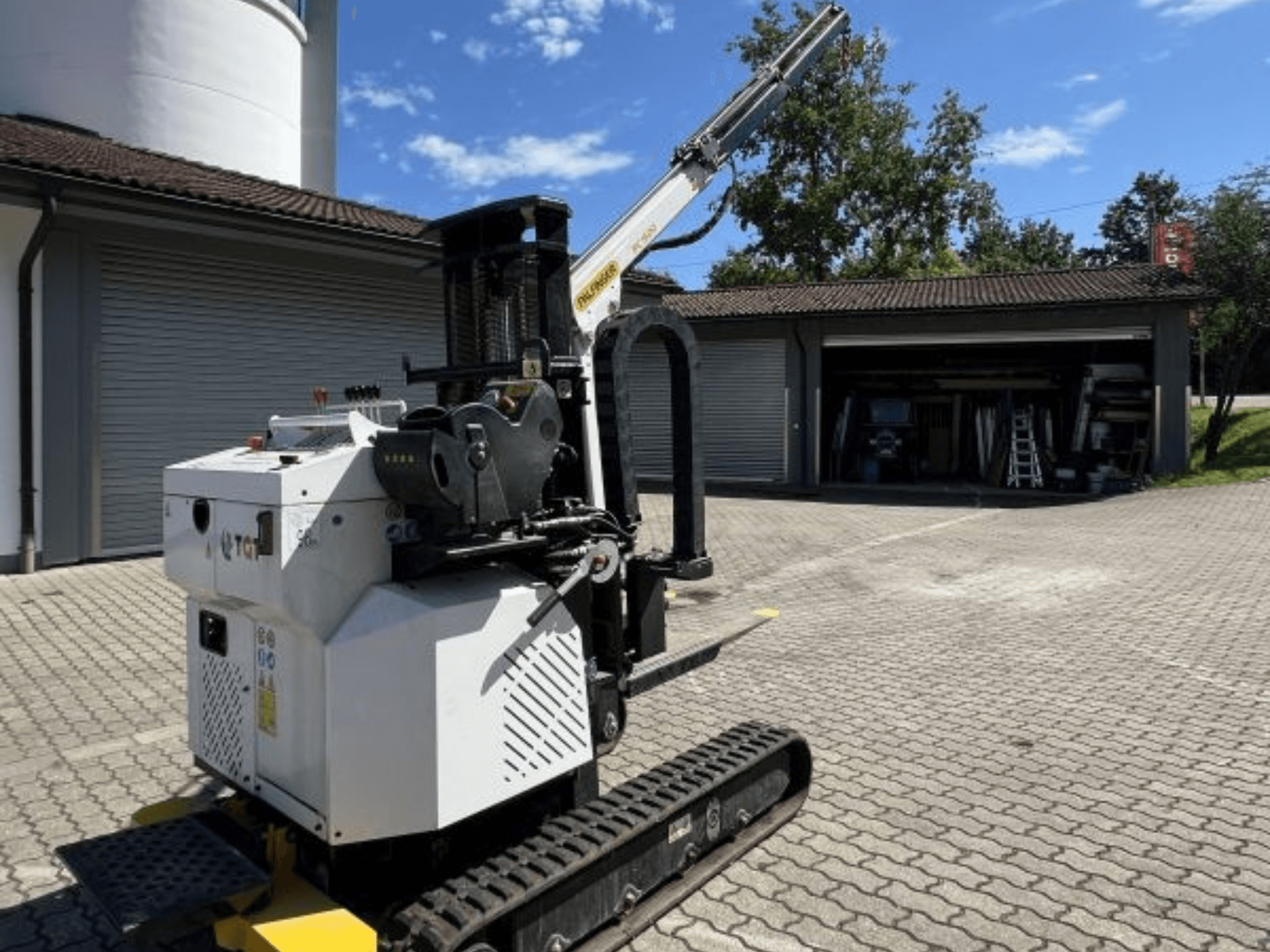 Stavební minijeřáb TGI, boční pohled na pásy a namontovaný výložník, zaparkovaný na cihlovém povrchu u garáže.