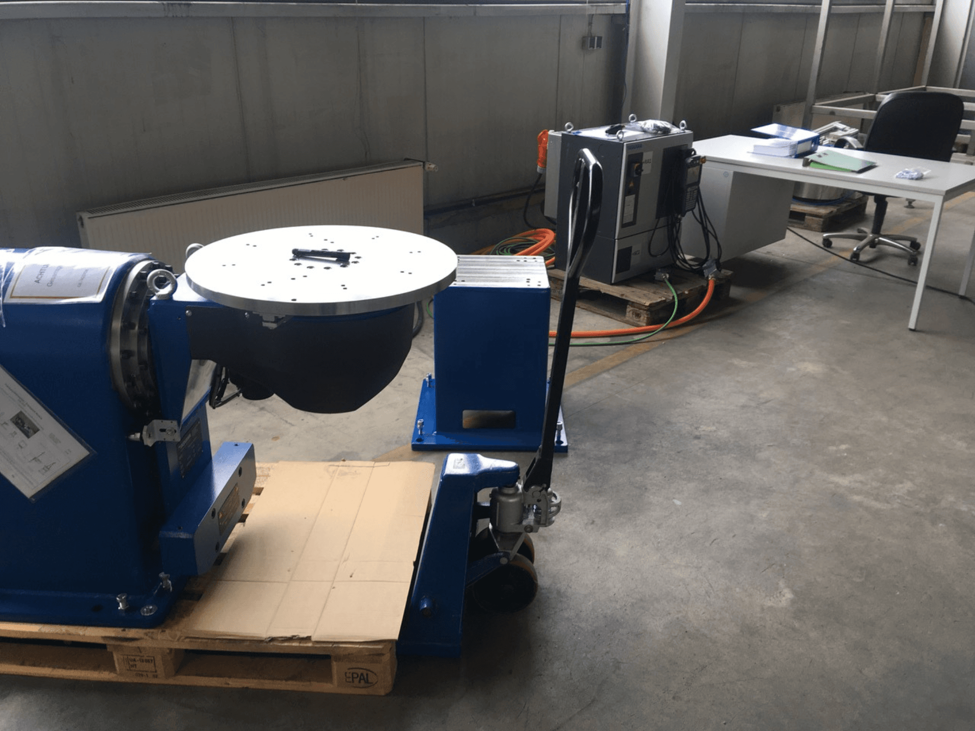 Modré průmyslové zařízení s kruhovou základnou a ovládacím panelem, pohled zepředu, na dřevěné paletě v kancelářském prostředí.