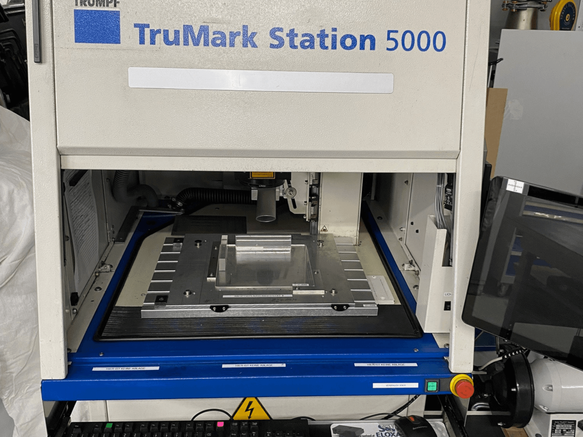 Pohled zepředu na laserový značkovací stroj TruMark Station 5000 s pracovním stolem, ovládacími prvky a monitorovací obrazovkou.