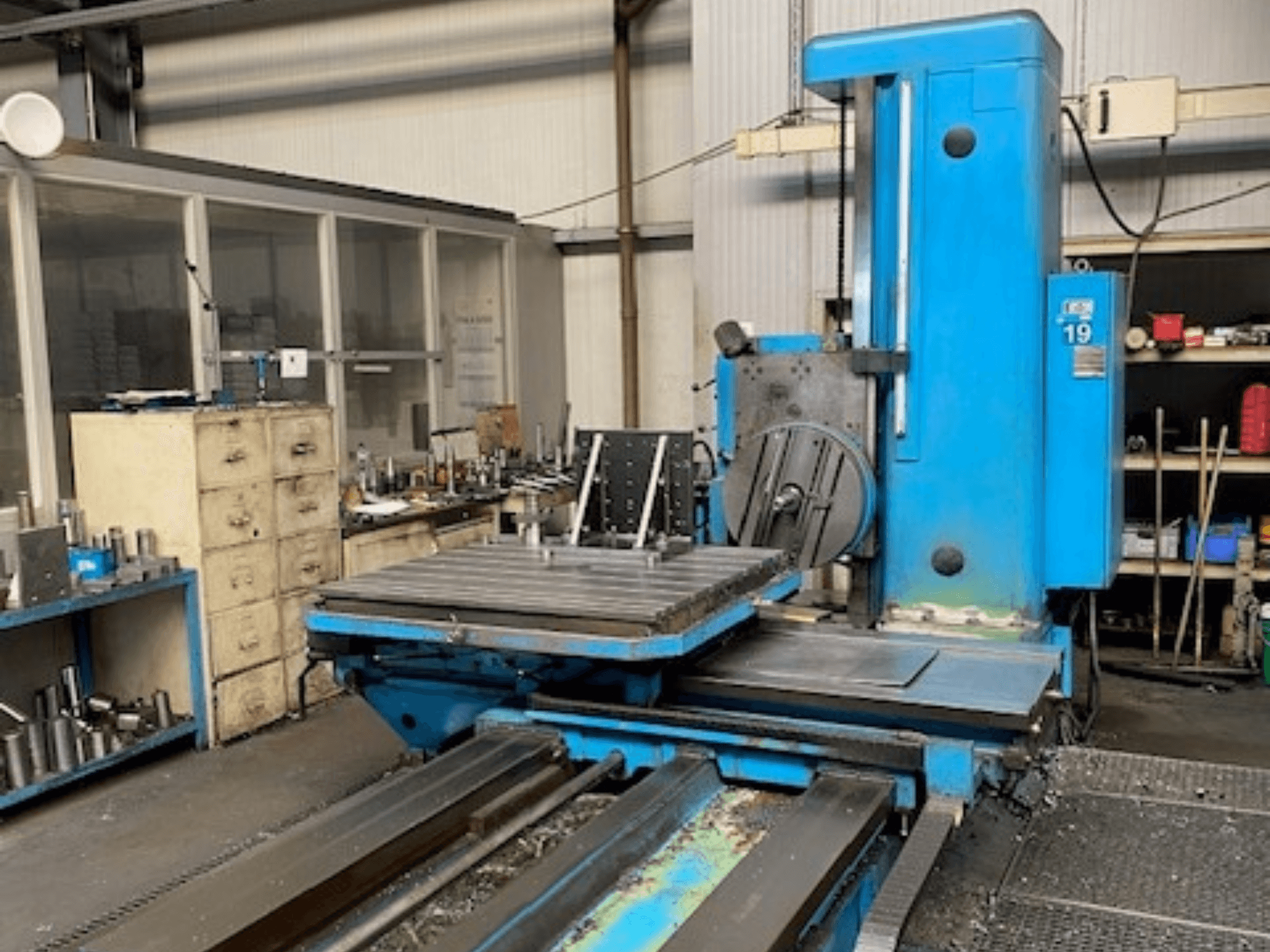 Modrá frézka v čelním pohledu, model číslo 19, na podlaze dílny s nářadím a úložnou skříní v pozadí.