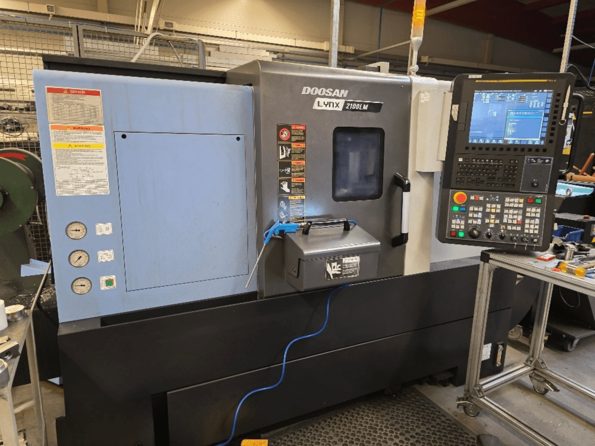 CNC soustruh Doosan Lynx 2100LM, pohled zepředu, s ovládacím panelem, manometry a bezpečnostními upozorněními.