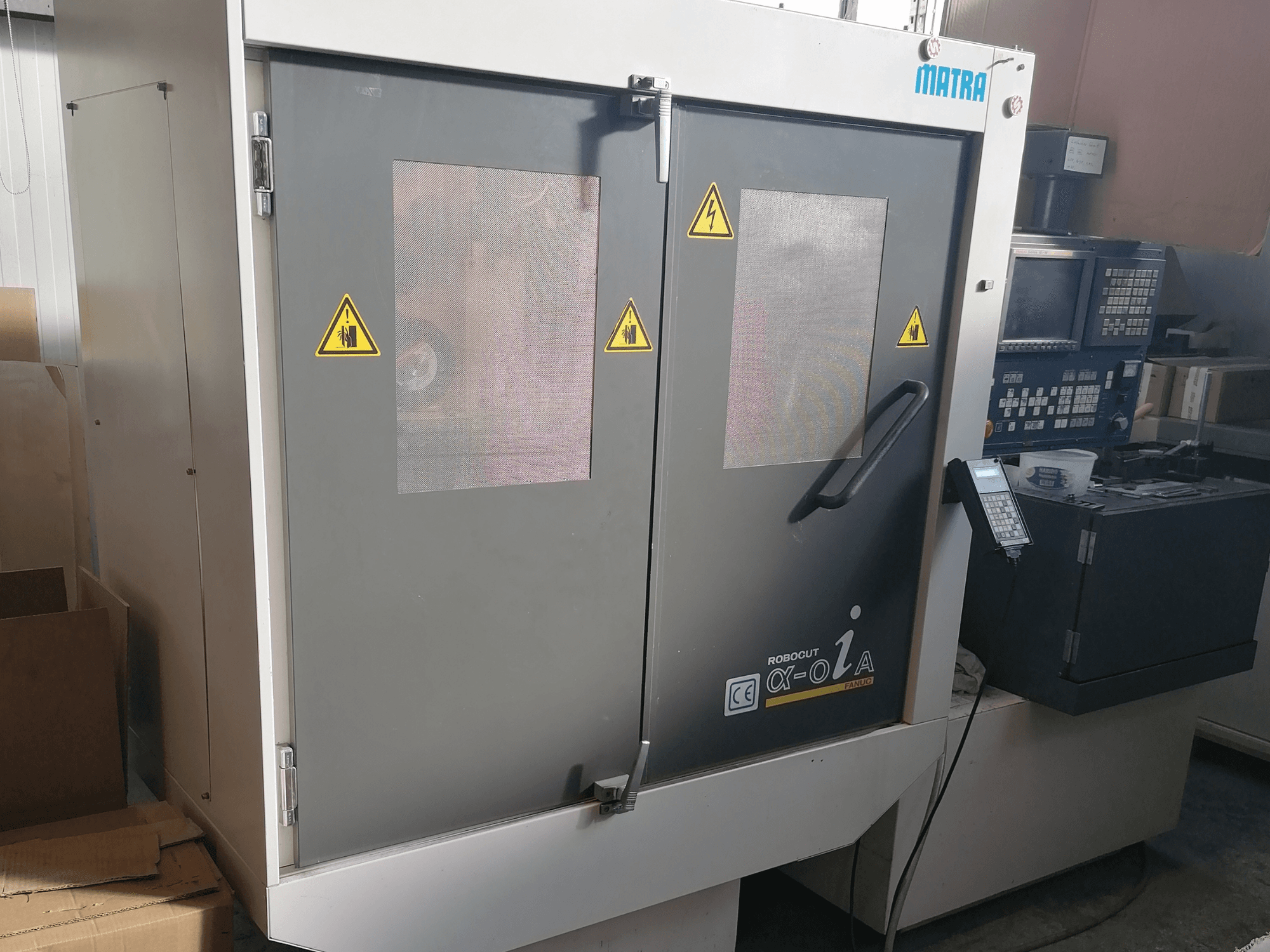 Stroj Robocut α-oZα od společnosti Matra, pohled zepředu s bezpečnostními upozorněními, ovládacím panelem a dvojitými dveřmi se síťovými okny.