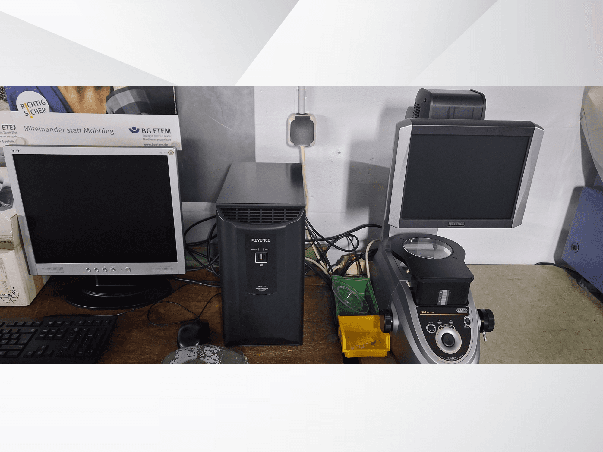 Dva počítačové monitory, jeden Acer a jeden Keyence, vedle černého zařízení a mikroskopu, pohled zepředu.