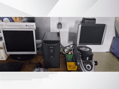 Dva počítačové monitory, jeden Acer a jeden Keyence, vedle černého zařízení a mikroskopu, pohled zepředu.