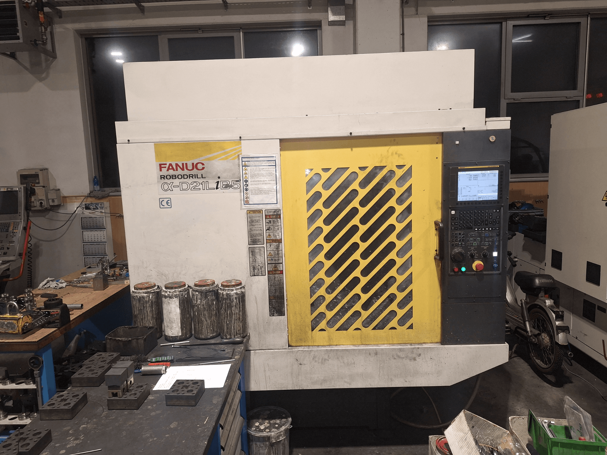 Pohled zepředu na vrtačku FANUC Robodrill α-D21LiB5, zobrazující ovládací panel, žlutou mřížku a blízké nástroje na pracovním stole.