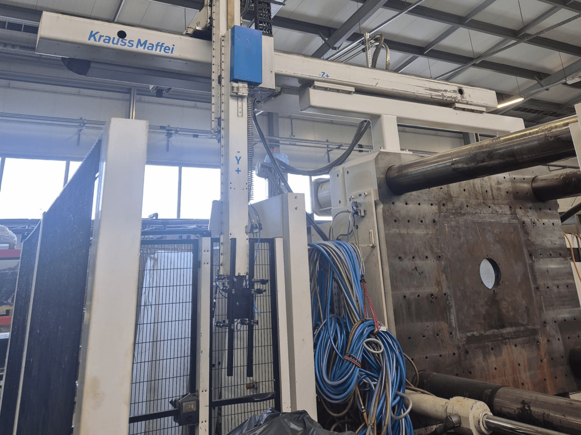 Hydraulický vstřikovací lis Krauss Maffei 1300-12000 MX, pohled zepředu s mechanickými rameny a hydraulickými přípojkami.