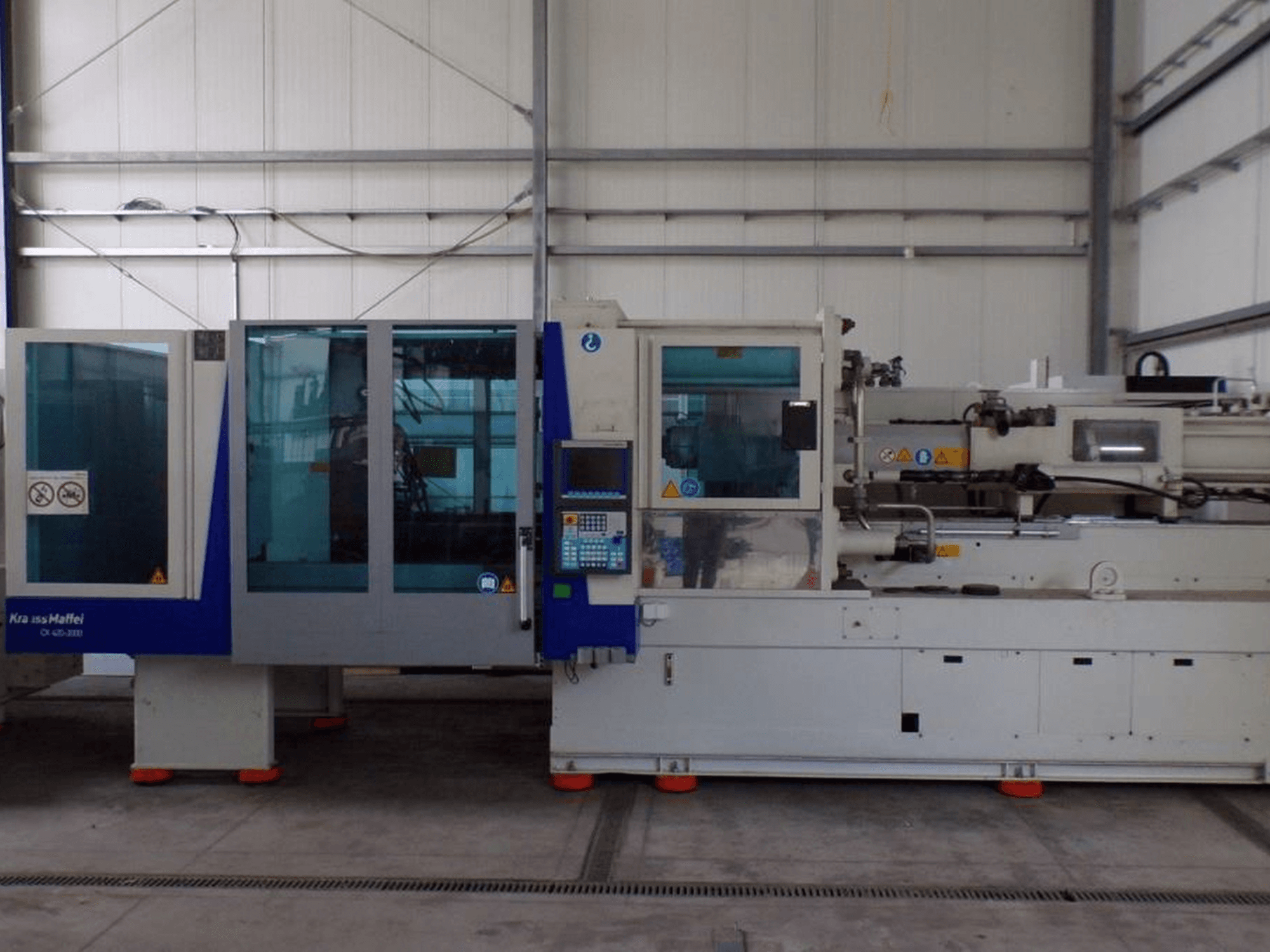 Vstřikovací stroj KraussMaffei, boční pohled, s ovládacím panelem a bezpečnostními ukazateli, viditelný v továrním prostředí.