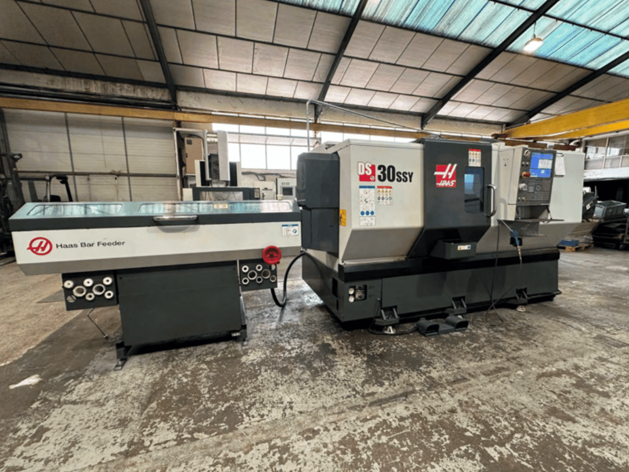 CNC soustruh Haas DS-30SSY s podavačem tyčí Haas, boční pohled na ovládací panel a podávací mechanismus v dílenském prostředí.