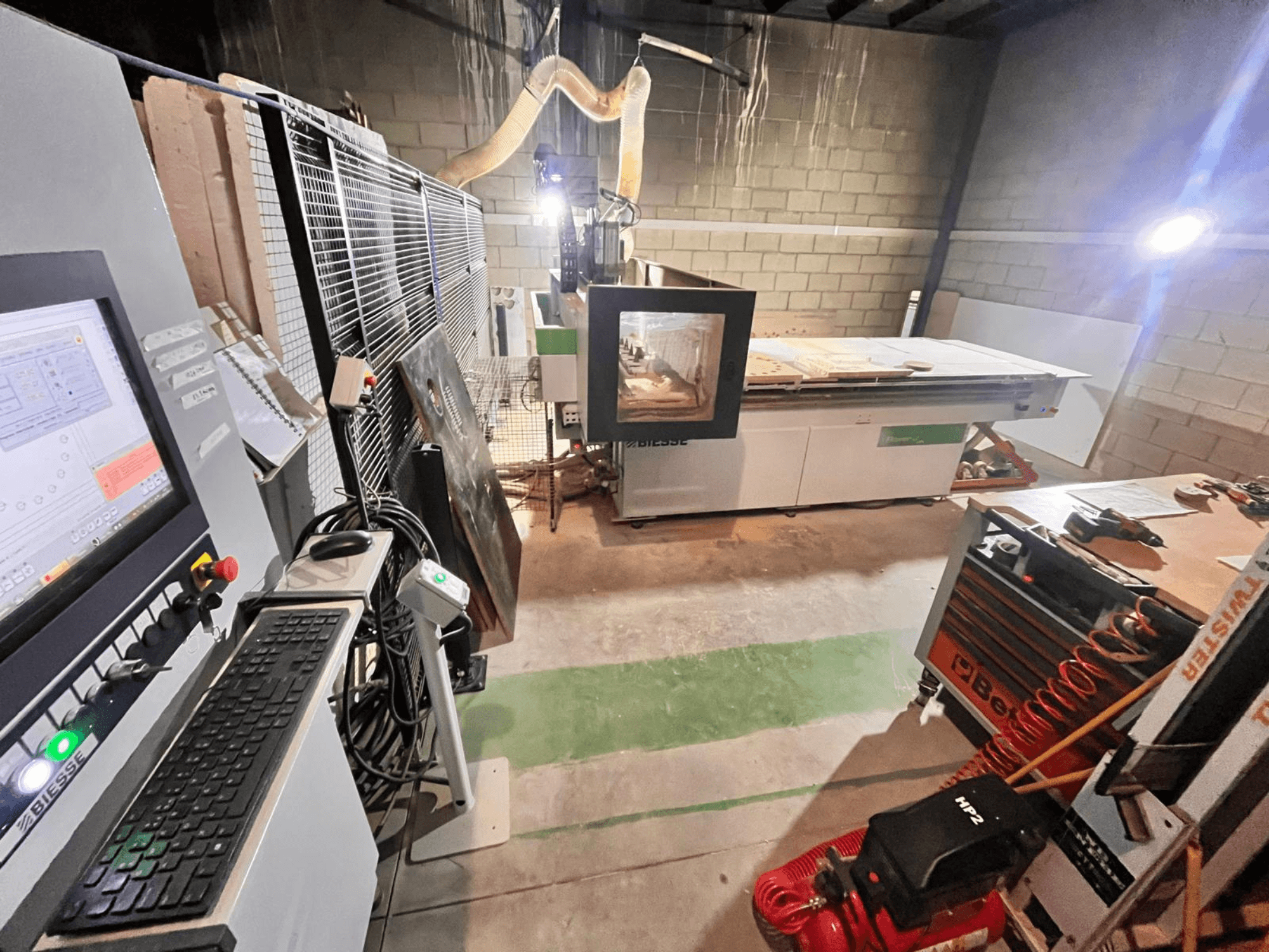 CNC stroj Biesse v dílně, boční pohled ukazuje ovládací panel, odsávání prachu, nástroje a různé materiály na stole.