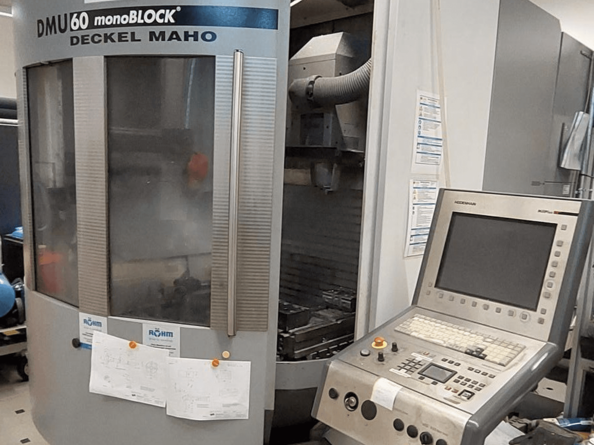 DMU 60 monoBLOCK společnosti Deckel Maho, čelní pohled na CNC frézku s ovládacím panelem a přiloženými technickými výkresy.