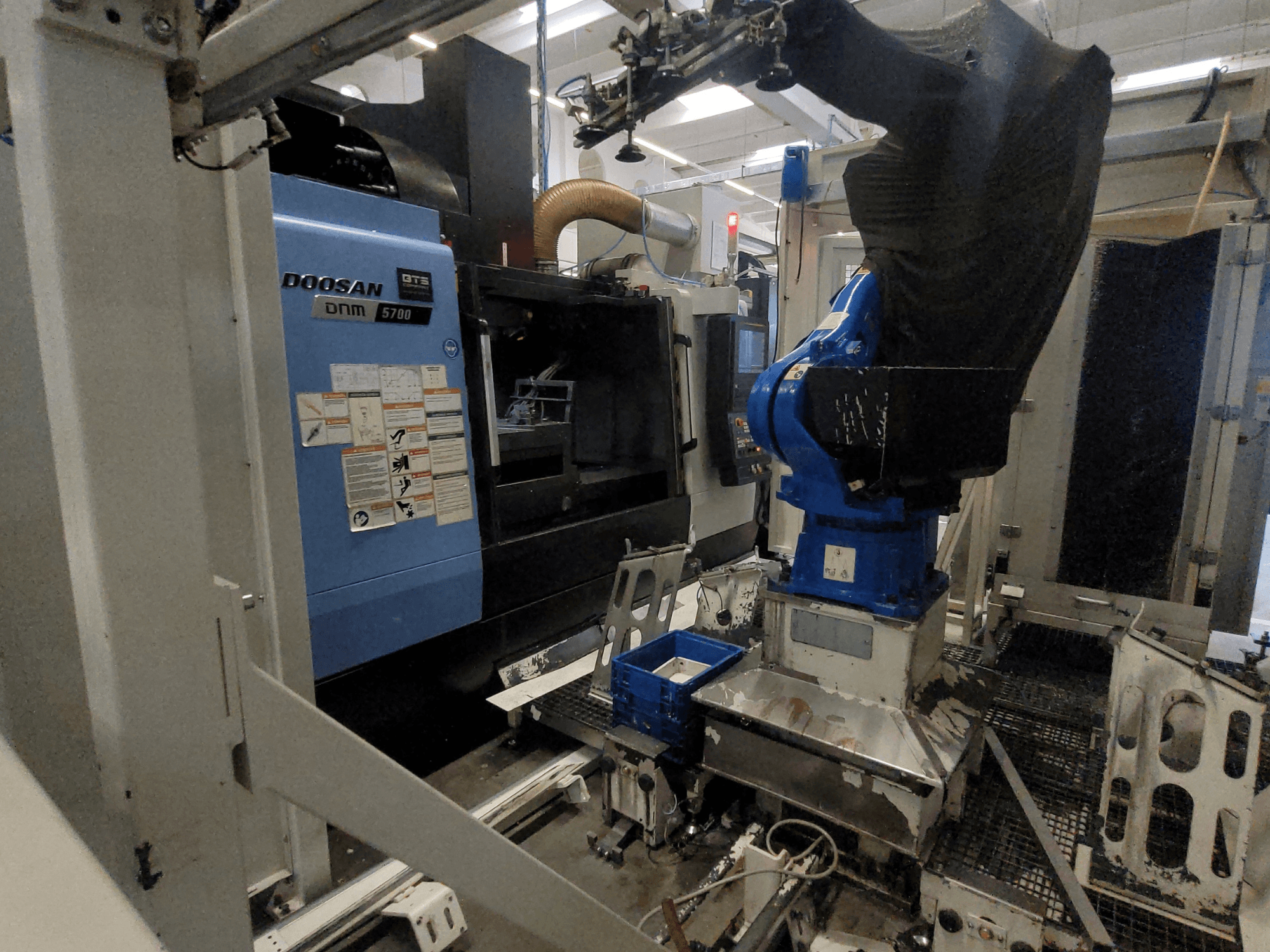 Vertikální obráběcí centrum DOOSAN DNM 5700 v továrním prostředí, na snímku je vidět jeho čelní pohled a integrace robotického ramene.