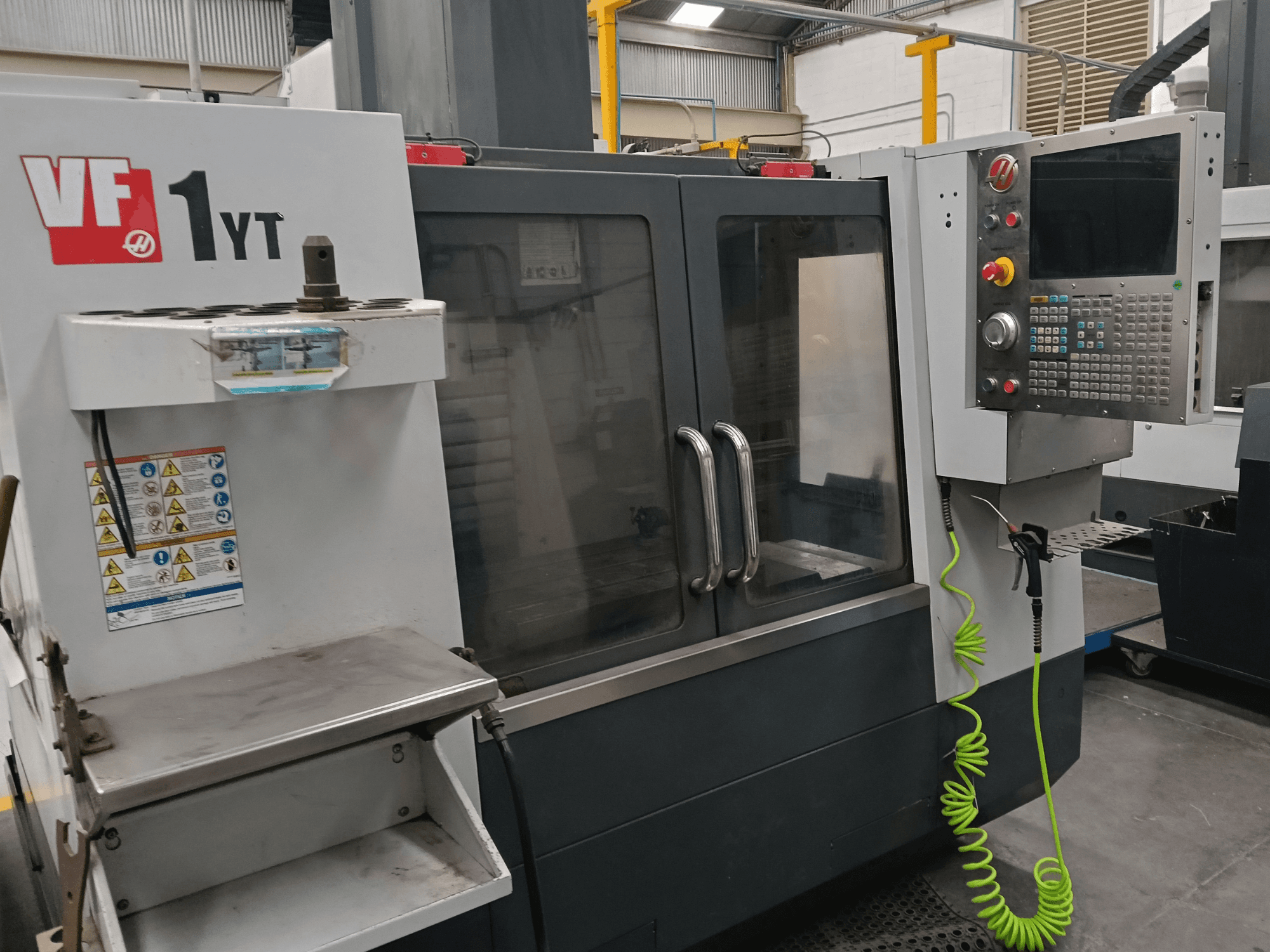Vertikální obráběcí centrum HAAS VF-1YT-2, pohled zepředu, na kterém je vidět ovládací panel, držák nástroje, bezpečnostní štítky a obráběcí prostor.