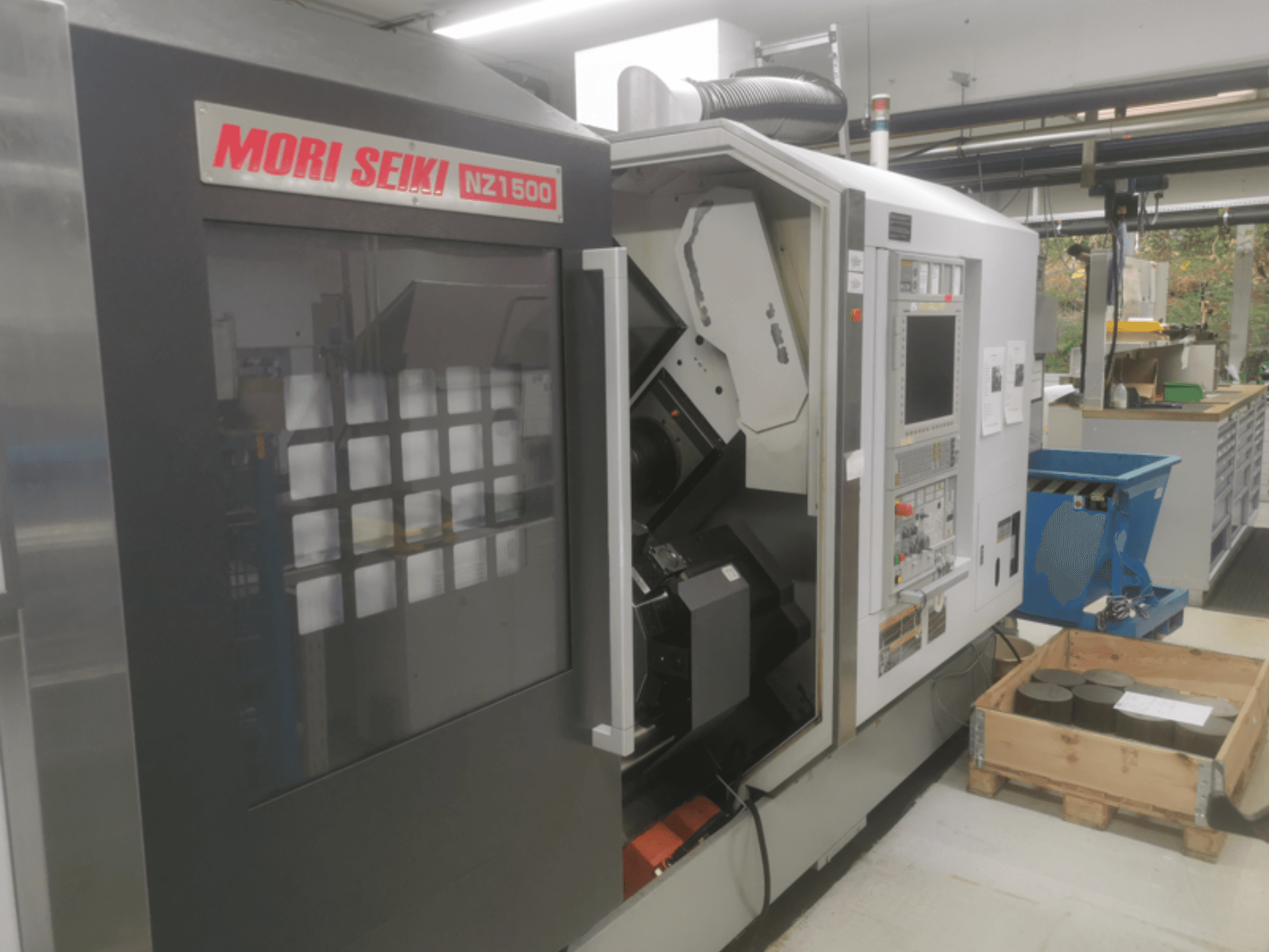 CNC stroj MORI SEIKI NZ1500 v mírném úhlu, ukazuje ovládací panel a pracovní prostor s kovovými obrobky na dřevěné paletě.