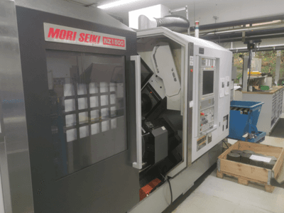CNC stroj MORI SEIKI NZ1500 v mírném úhlu, ukazuje ovládací panel a pracovní prostor s kovovými obrobky na dřevěné paletě.
