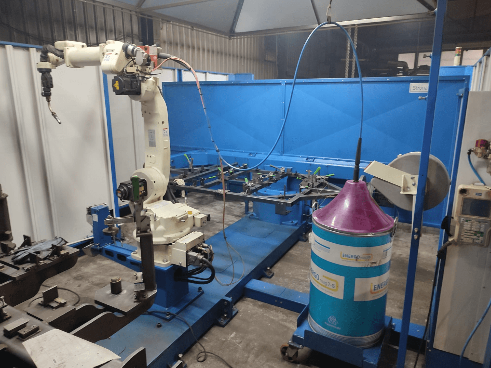 Průmyslové robotické rameno s bílou a modrou základnou, nastavené v dílně s modrou pracovní plochou a kovovými součástmi.